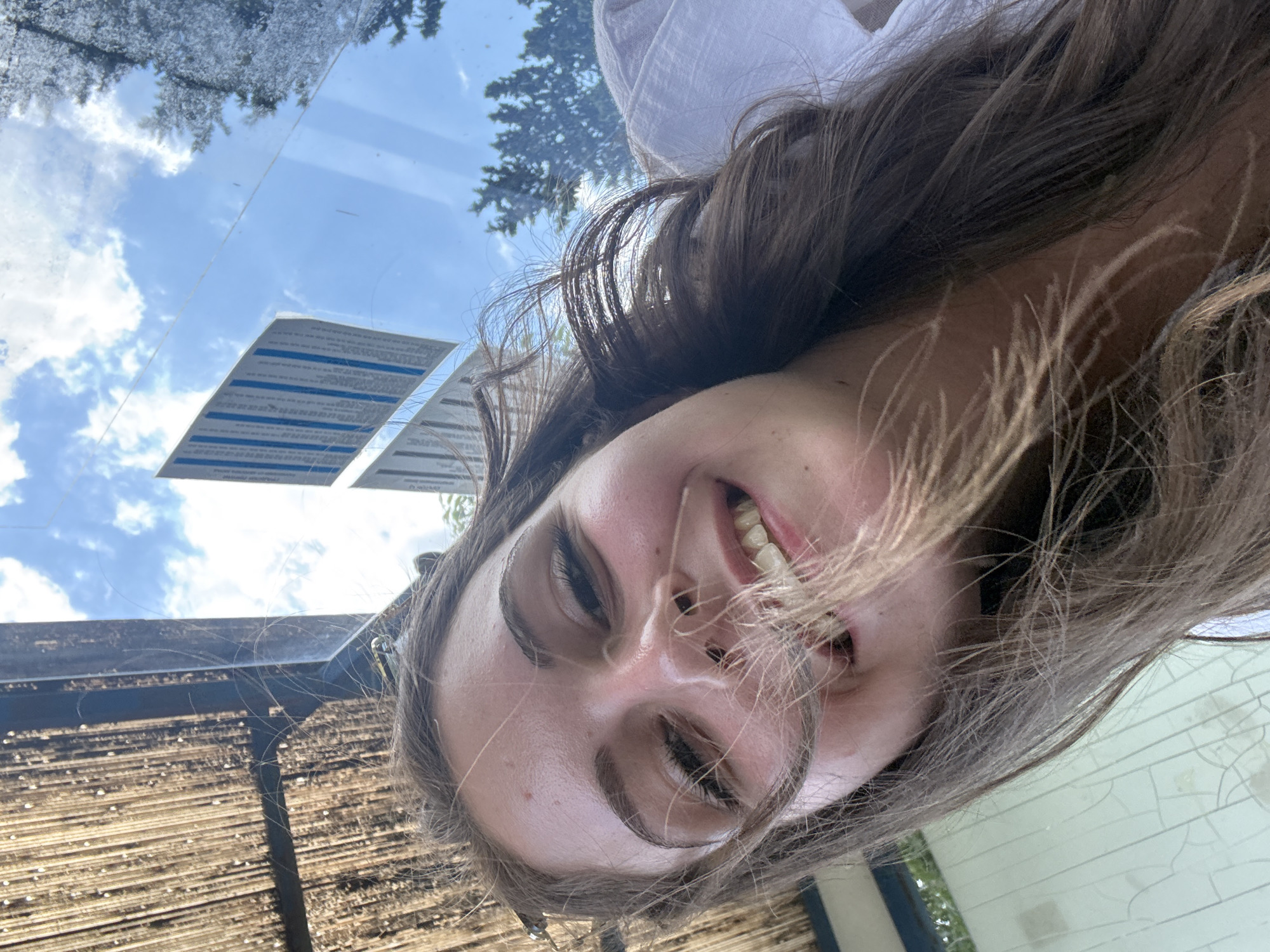 A young woman with long, brown hair smiles broadly at the camera in a selfie. Her hair is being blown by the wind across her face. She is wearing a white top. In the background, there is a thatched roof structure, a clear sky with clouds, and trees. A laminated notice is visible on a glass surface. The image appears to be taken in an outdoor setting. The location is identified as Burgas, Bulgaria, likely a public area given the notice and sky visible.