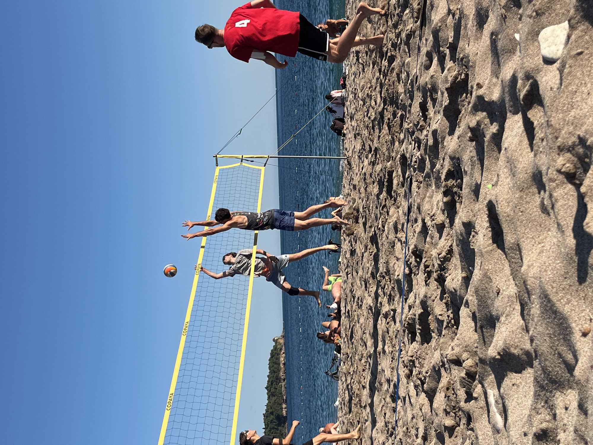 In Zankovići, Montenegro, a beach volleyball game is underway on a sunny day. The scene captures four young men in action. On one side of the net, a player in a grey t-shirt and dark shorts is mid-air, reaching for the ball. His teammate, wearing a black tank top and blue shorts, is also jumping, his arms extended. Facing them across the net, a player in a red t-shirt with the number "4" on the back and black shorts watches the play, while his teammate, also in black shorts, is partially visible on the far left. The beach is sandy, with the textured surface of the sand prominently featured in the foreground. Beyond the volleyball court and the players, the turquoise waters of the Adriatic Sea stretch out, with a distant, tree-covered coastline visible under a clear blue sky. Several other beachgoers are scattered in the background, some relaxing on the sand.