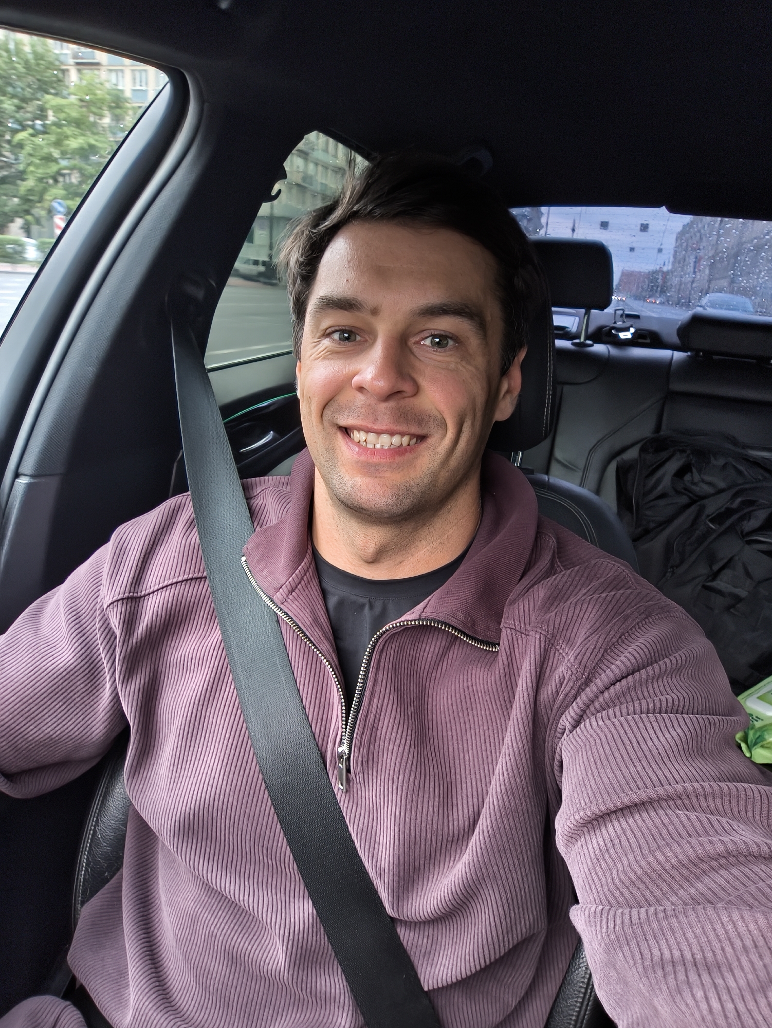 A man is in the driver's seat of a car, likely a BMW based on the interior details. He is wearing a purple corduroy jacket over a black t-shirt and a black seatbelt is fastened across his chest. The man is smiling and looking at the camera. The car appears to be on a city street with buildings and trees visible outside the windows. The background suggests a European city, possibly Saint Petersburg, Russia, with overcast skies and a hint of rain on the windows. The interior of the car is clean and modern, with black leather seats and a black headliner. A black jacket is visible on the back seat.