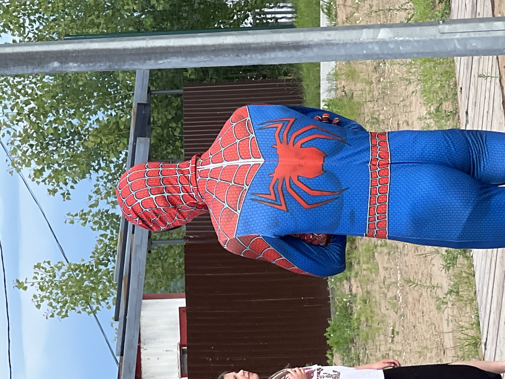 The image shows a person from behind wearing a blue and red Spider-Man costume, standing outdoors in Chaikovsky, Russia. The person is facing away from the camera, with their arms crossed. The costume features the iconic red web pattern on a blue background, with a large red spider emblem on the back. To the left, a young girl wearing a white t-shirt and a black skirt is partially visible, looking towards the Spider-Man character.
In the background, there is a brown wooden fence, a white building with a red trim, and a metal pole on the right side of the image. Green trees and foliage are visible behind the fence and building. The ground is covered with dirt, sparse vegetation, and a wooden walkway in the foreground. The sky is visible above, with some clouds. The scene appears to be in an outdoor recreational or park-like setting.
