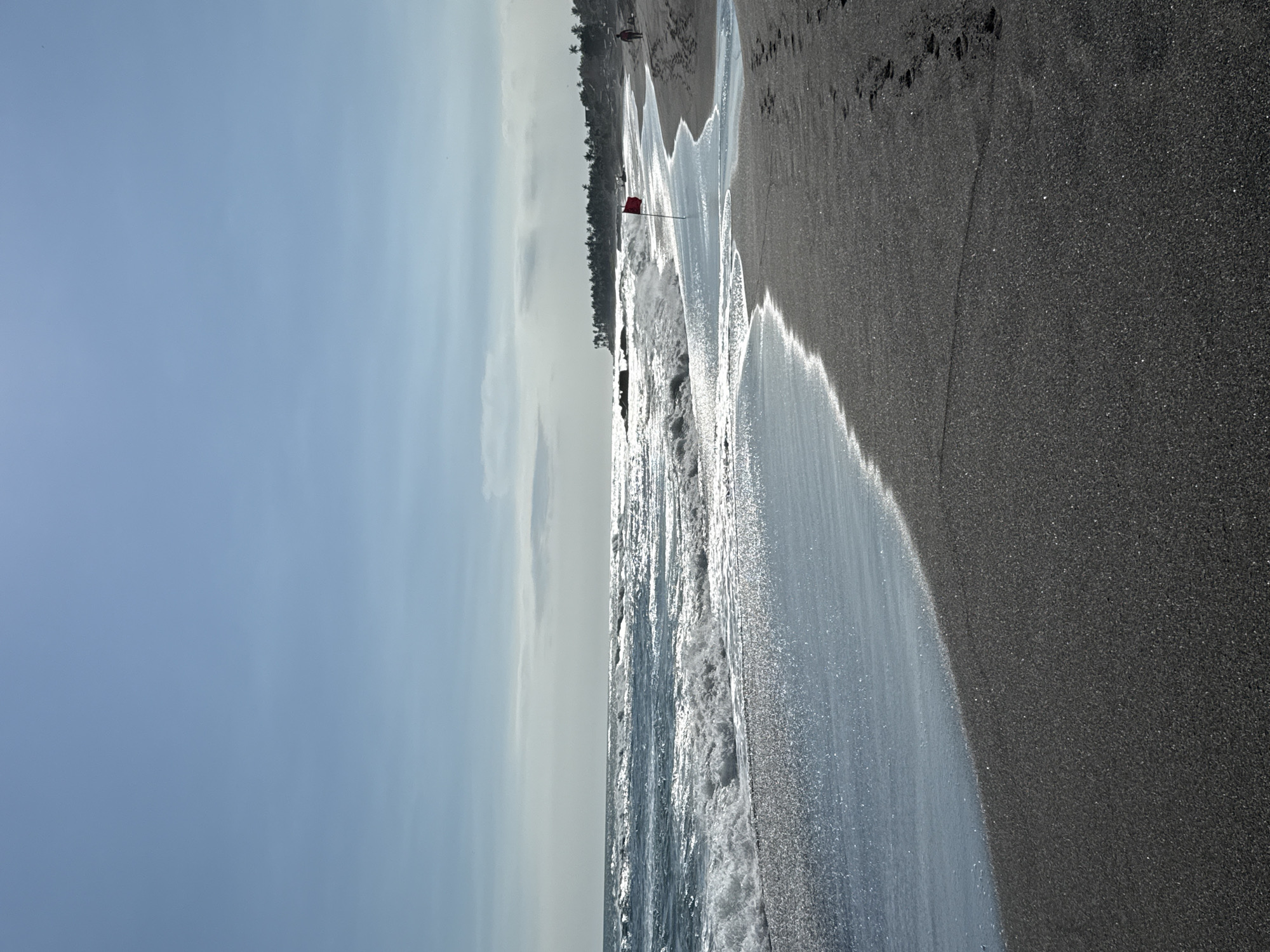 The image depicts a shoreline, likely in Mengwi, Indonesia, given the request's context. The beach has dark sand and is being washed by waves. A red flag is visible on the beach, likely a warning signal. The sky is blue, with some cloud cover. There is a figure walking away in the distant horizon line.