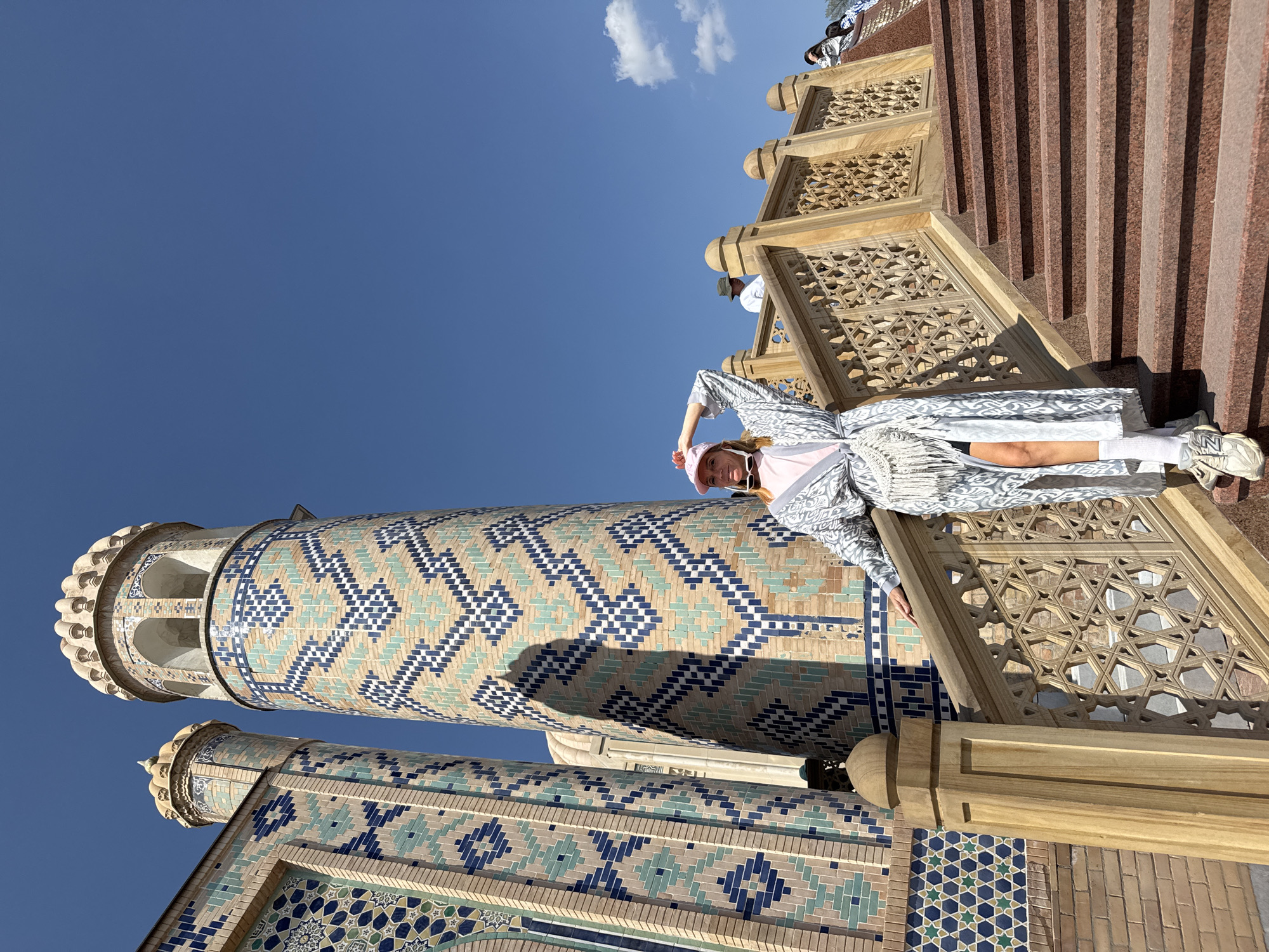 The image shows a woman in a pink hat, a light-colored patterned robe, and white socks, standing on carved wooden steps next to a tall, mosaic-covered minaret. The minaret features intricate geometric patterns in blue, turquoise, and beige. The sky is clear blue with some clouds. The location is likely a historical or religious site in Samarkand City, Uzbekistan, given the architectural style.