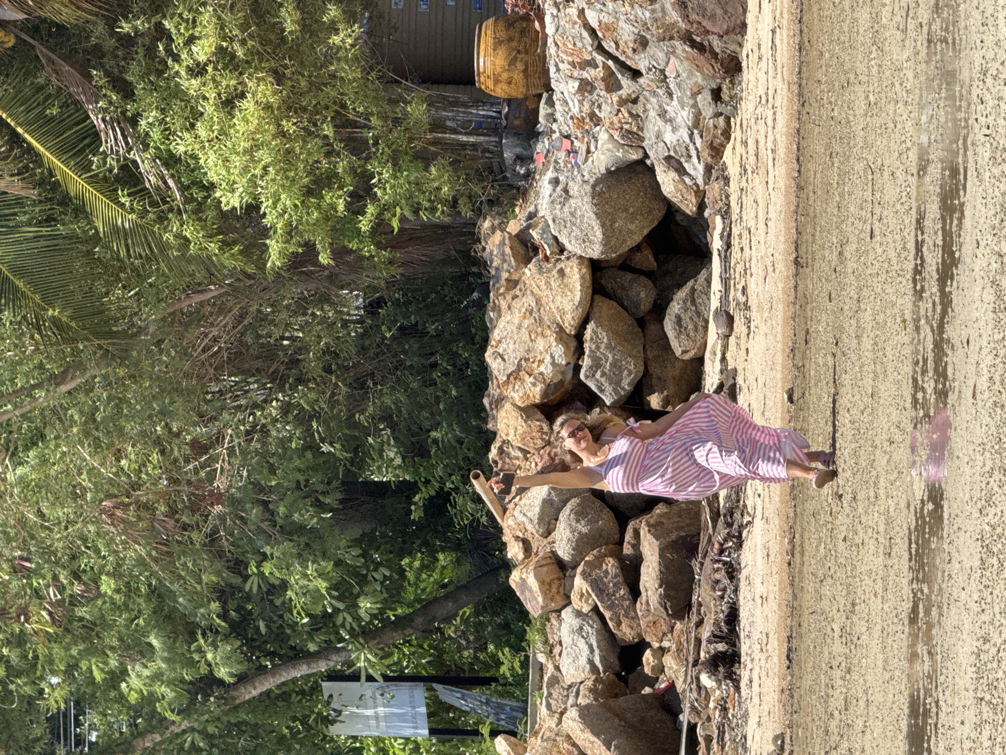 In a scene on a beach in ตำบลเกาะพะงัน, Thailand, a woman in a pink and white striped dress is captured mid-motion, holding a smartphone aloft in her right hand as if taking a selfie or recording a video. She is looking towards the camera, with a slight smile and sunglasses shielding her eyes. The woman stands on a sandy beach, with a shallow pool of water reflecting the light in the foreground. Behind her, a large pile of rocks forms a natural barrier, with lush green foliage and trees growing behind the rocks, suggesting a tropical environment. A signpost is partially visible on the left side of the image. The overall impression is a sunny day at a coastal location.