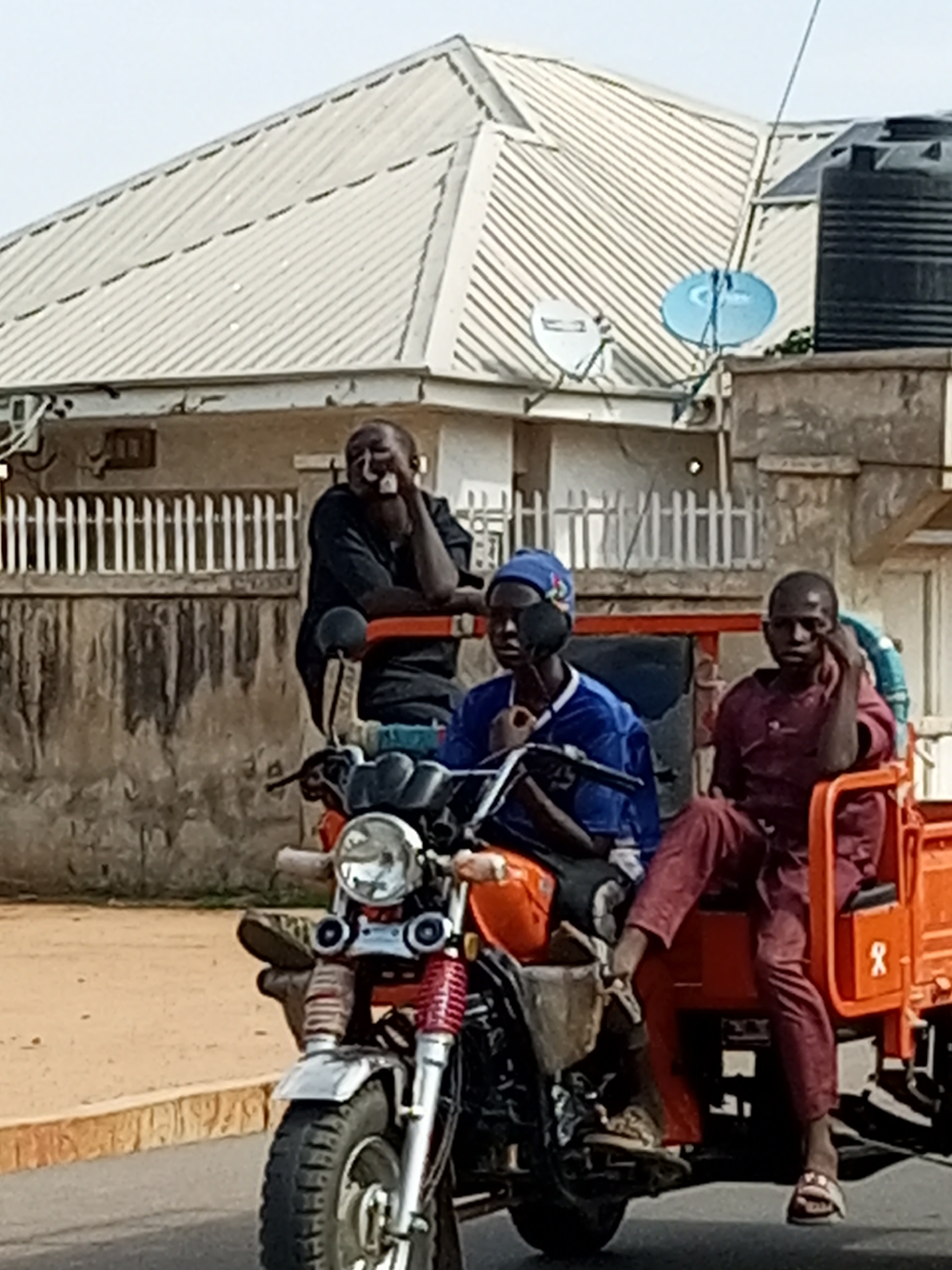In Gombe, Nigeria, on a paved road, a three-wheeled motorcycle with an orange cargo bed is being ridden by three young men. The rider in the front is wearing a blue headscarf and a blue and white jersey. The middle passenger, seated behind the rider, is wearing a dark shirt and leaning his head on his hand. The passenger on the right wears a burgundy outfit and is also resting his head on his hand. Behind them, a building with a corrugated metal roof, possibly a home or business, is visible. The building has white railings in front and a large black water tank on the right side. Two satellite dishes are mounted on the roof of the building, one blue and one white. The men appear relaxed and are likely commuting or traveling within the city.