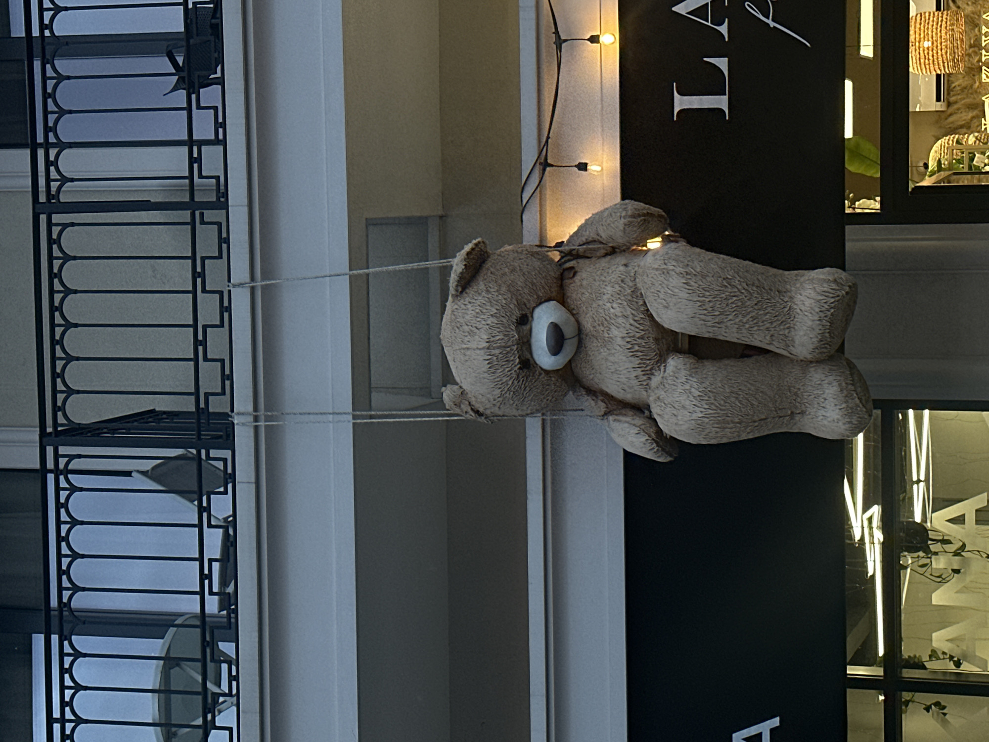 A large, light brown teddy bear, appearing to be a decorative fixture, hangs suspended by ropes in front of a building in Budva, Montenegro. The bear is positioned in a sitting posture, facing forward, with its white muzzle and dark nose prominent.
Above the bear, a balcony features a black wrought iron railing with curved and straight vertical bars, connected by a lower horizontal patterned section. Behind the railing, a round light-colored table and at least one matching chair are visible on the balcony, alongside a large window or glass door. The building facade at this level is a light, neutral color. One of the suspension ropes for the teddy bear extends downwards from the balcony railing.
Below the balcony, a string of decorative lights with exposed bulbs is strung horizontally across a dark-colored commercial fascia board. Partial white lettering is visible on this fascia: "ANA" on the left and "LA" and a script "f" on the right. Below the fascia, a storefront with large glass windows is visible. Reflections in the left window show linear light fixtures and the repeated letters "ANA." In the right storefront window, an illuminated woven lampshade and green foliage are discernible inside the establishment. No people are visible in the image. The ambient lighting suggests dusk or nighttime.