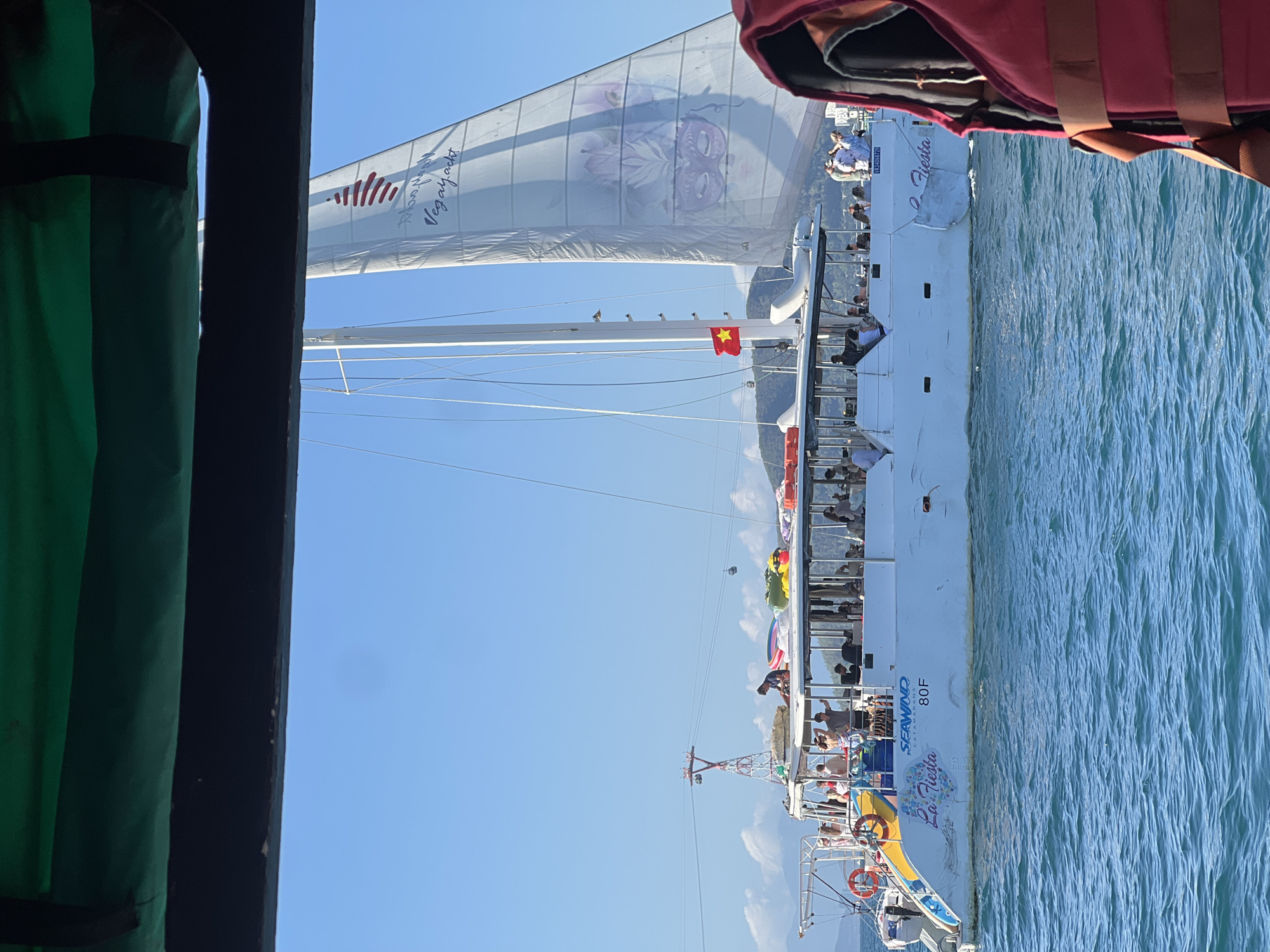 The image shows a view from a boat in what appears to be Nha Trang Ward, Vietnam. A large catamaran named "La Fiesta," specifically a "SEAWIND Catamaran 80F," is visible alongside. The boat is carrying numerous passengers. The sail of a neighboring catamaran has "vegan.yacht" written on it and a red striped logo. A Vietnamese flag is flying on the catamaran. Part of a person wearing a red hooded jacket and a life vest is visible in the upper right corner. To the left, there is a dark structure with a green curtain. The sea is visible, and in the background, there are mountains or large hills. The sky is clear and blue.