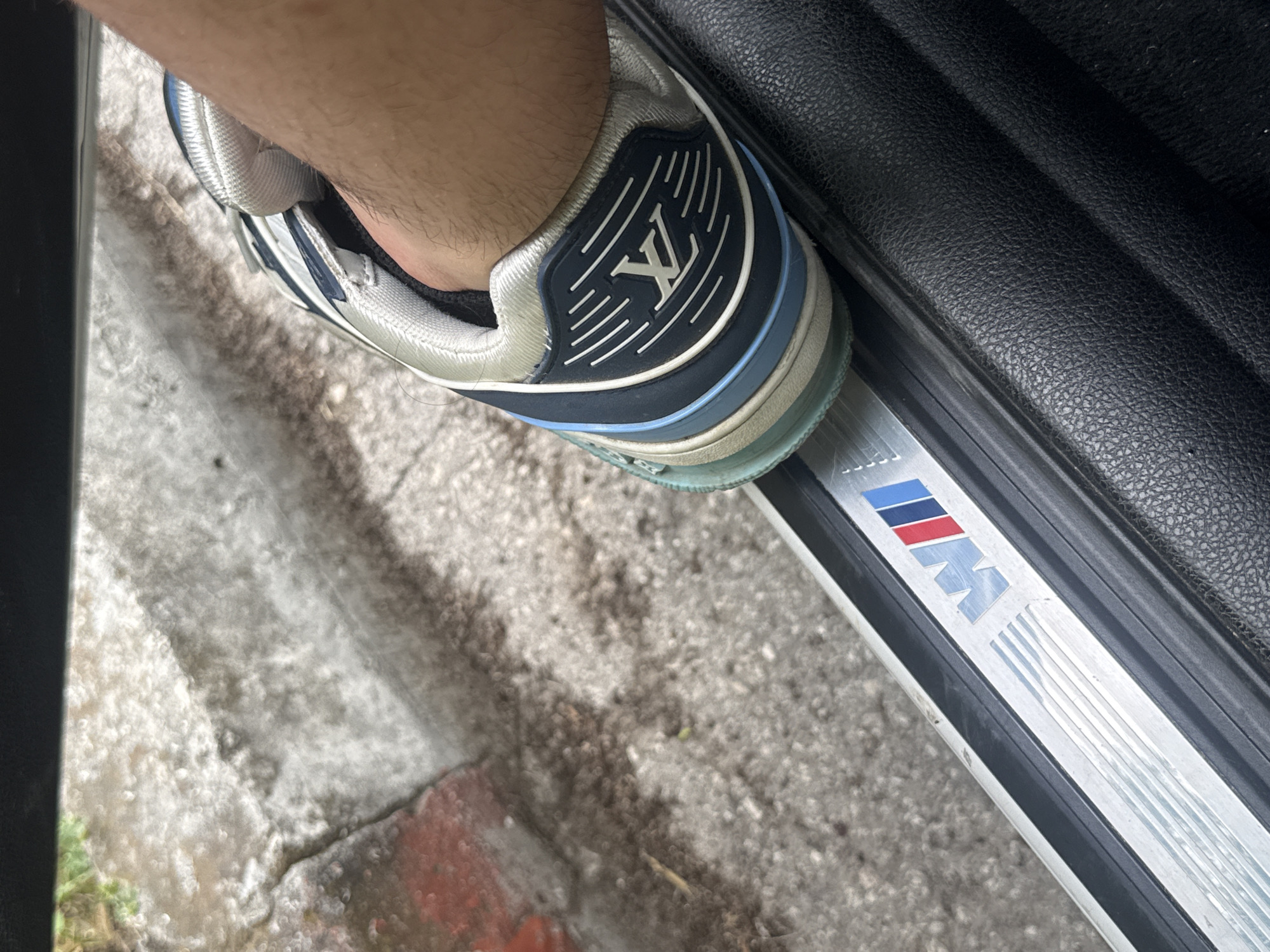 A partial view of a person's foot wearing a blue and white Louis Vuitton sneaker is resting on a silver BMW car door sill. The car door sill has the "M" logo in red, blue, and white. The background shows a concrete surface and a portion of a black car interior. The location appears to be outdoors, based on the concrete. The city is not discernible from the image.