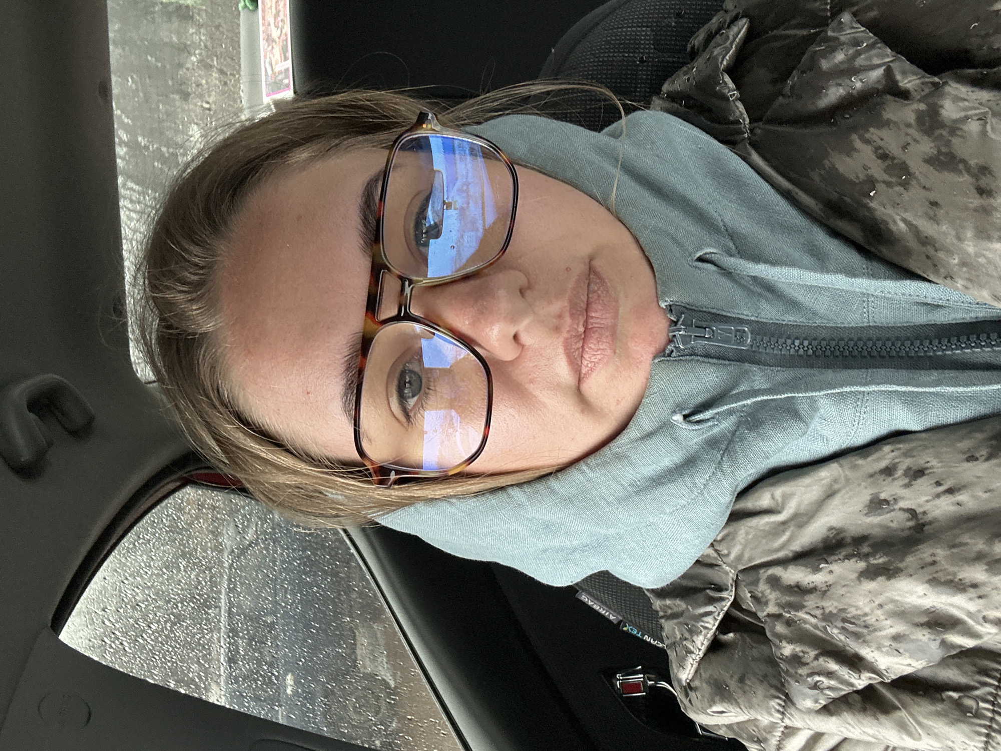 The image shows a person wearing glasses and a hooded jacket inside a car. The car window is wet, suggesting it is raining. The location is indeterminate. Ural'skoe, Russia, cannot be confirmed.