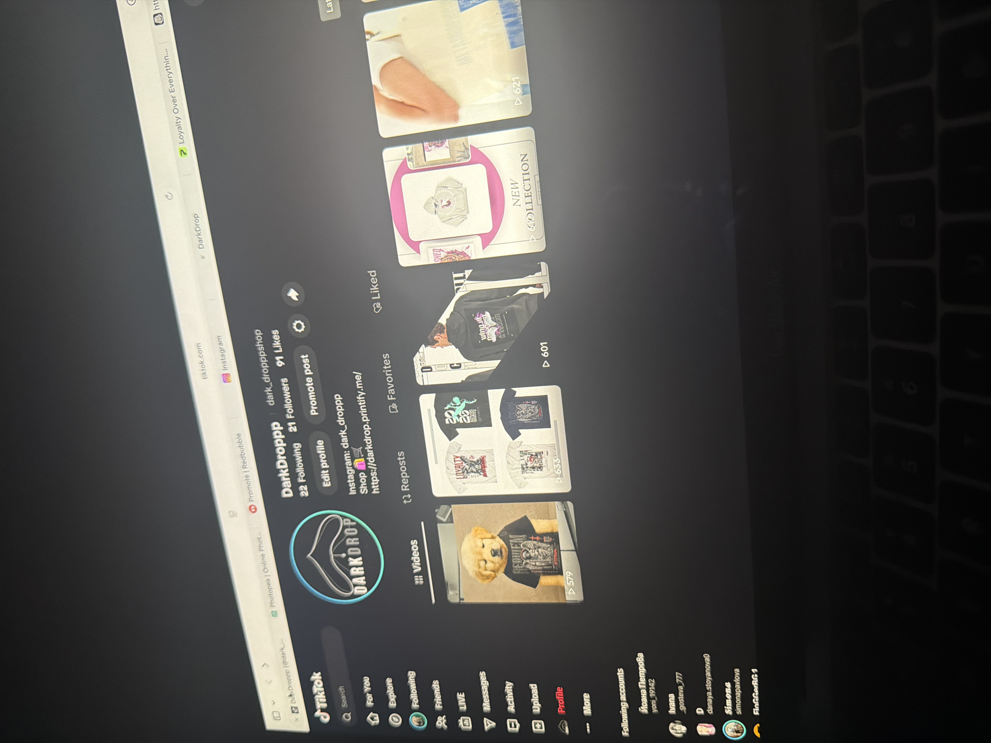 A laptop screen, angled slightly away from the viewer and illuminated against a dark background, displays the TikTok profile for "DarkDroppp" (@dark_dropppshop). The screen shows several open browser tabs at the top, including "tiktok.com," "Photopea | Online Phot...," "Promote | Redbubble," "Instagram," "DarkDrop," and "Loyalty Over Everythin..."
The TikTok interface features a left sidebar with navigation options such as "For You," "Explore," "Following," "Friends," "LIVE," "Messages," "Activity," "Upload," and "Profile," which is highlighted in red. Below these, a "Following accounts" list is partially visible, showing names like "Йоана Петрова (yoni_19142)," "Ivana (_gosteva_777)," "D (danaya.stoyanova0)," "Simone (simonapavlova)," and "BloGGeRGB 1," indicating a connection to Bulgaria.
The main content area presents the "DarkDroppp" profile, featuring a circular logo with a stylized droplet shape and the brand name "DARKDROP." Profile statistics display "22 Following," "21 Followers," and "91 Likes." Below this, "Edit profile" and "Promote post" buttons are visible. The profile bio includes "Instagram: dark_droppp" and a shop link: "https://darkdrop.printify.me/."
The content is categorized into "Videos," "Reposts," "Favorites," and "Liked," with "Videos" currently selected. Five video thumbnails are displayed:
1. A golden retriever puppy wearing a black t-shirt with "REQUIEM ETERNAL" text and a gothic graphic, with 579 views.
2. A collage of four different t-shirt designs, one with "LOYALTY" text and another with "TEMPO + FLIP" text, accumulating 633 views.
3. A person wearing a black hoodie with a white graphic and partially visible text, "wing of freedom," viewed 601 times.
4. A promotional image for a "NEW COLLECTION," featuring a light-colored hoodie with a pink flamingo graphic, indicated by pink arrows and a "SHOP NOW" prompt.
5. A person holding a light-colored bag, possibly a tote, with 621 views.
The lower portion of the image reveals part of the laptop's black keyboard, with visible keys including "5," "6," "7," and "8." The ambient lighting is dark, with the laptop screen serving as the primary light source, and faint dust or reflections are present on the upper part of the screen. The scene is situated in a dark environment within Plovdiv, Bulgaria, evidenced by the Bulgarian names and handles visible on the TikTok profile.