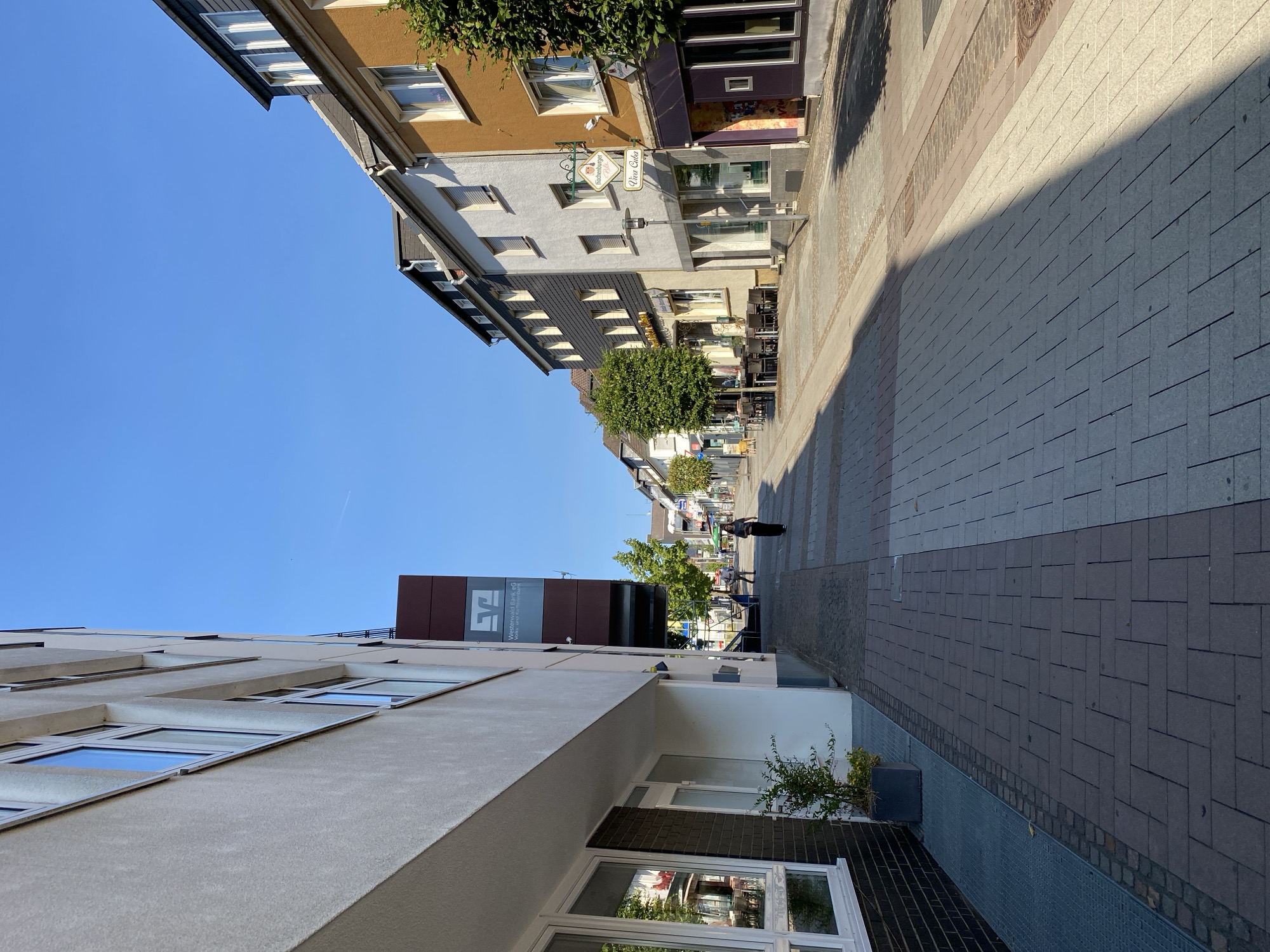 The image captures a street scene in Altenkirchen, Germany, under a clear blue sky. The perspective is from a pedestrian's viewpoint, looking down a cobblestone street lined with buildings.
On the left, a modern building with a light-colored facade dominates the foreground, featuring large windows and a section of dark brickwork. A sign for "Westerwald Bank" is visible on a prominent burgundy structure attached to this building.
The street itself is paved with interlocking cobblestones in varying shades of grey and beige, with a clear division of pedestrian walkways and a lighter area in the center. Outdoor seating with tables and chairs is set up in front of some of the establishments.
The buildings on the right display a mix of architectural styles and colors. One building is a deep orange, another is white, and further down, there are darker facades. Signs indicate businesses, including one with "Röschenburger" and "Vito Cara" on it, suggesting a restaurant or café.
A few individuals are present on the street. One person is walking down the center of the street, and another can be seen further in the distance. Trees with green foliage are interspersed along the street, providing some shade. The overall impression is a well-maintained, sunny street in a German town.
