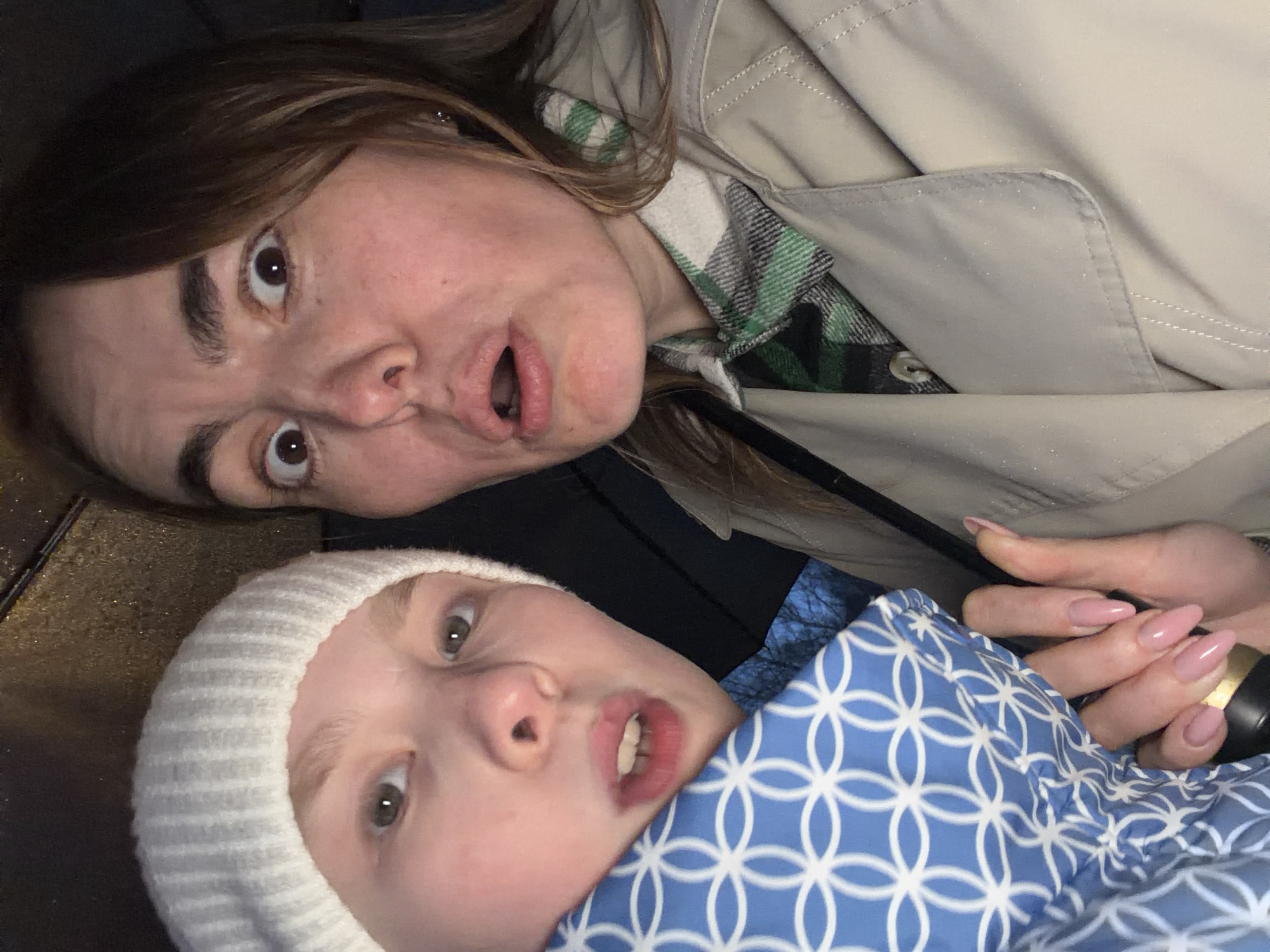 A selfie-style image shows a woman and a child, both with surprised expressions and open mouths. The woman, positioned at the top, has brown hair and is wearing a tan trench coat over a green and black plaid shirt. The child, wearing a white knitted hat and covered by a blue blanket with a white geometric pattern, is located at the bottom of the frame. The woman's hands, with light pink manicured nails, are holding a black object (possibly part of an umbrella) near the blanket. The background is dark, and there are reflections suggestive of wet pavement.
There is insufficient information in the image to determine the location in the city of Izhevsk, Russia.