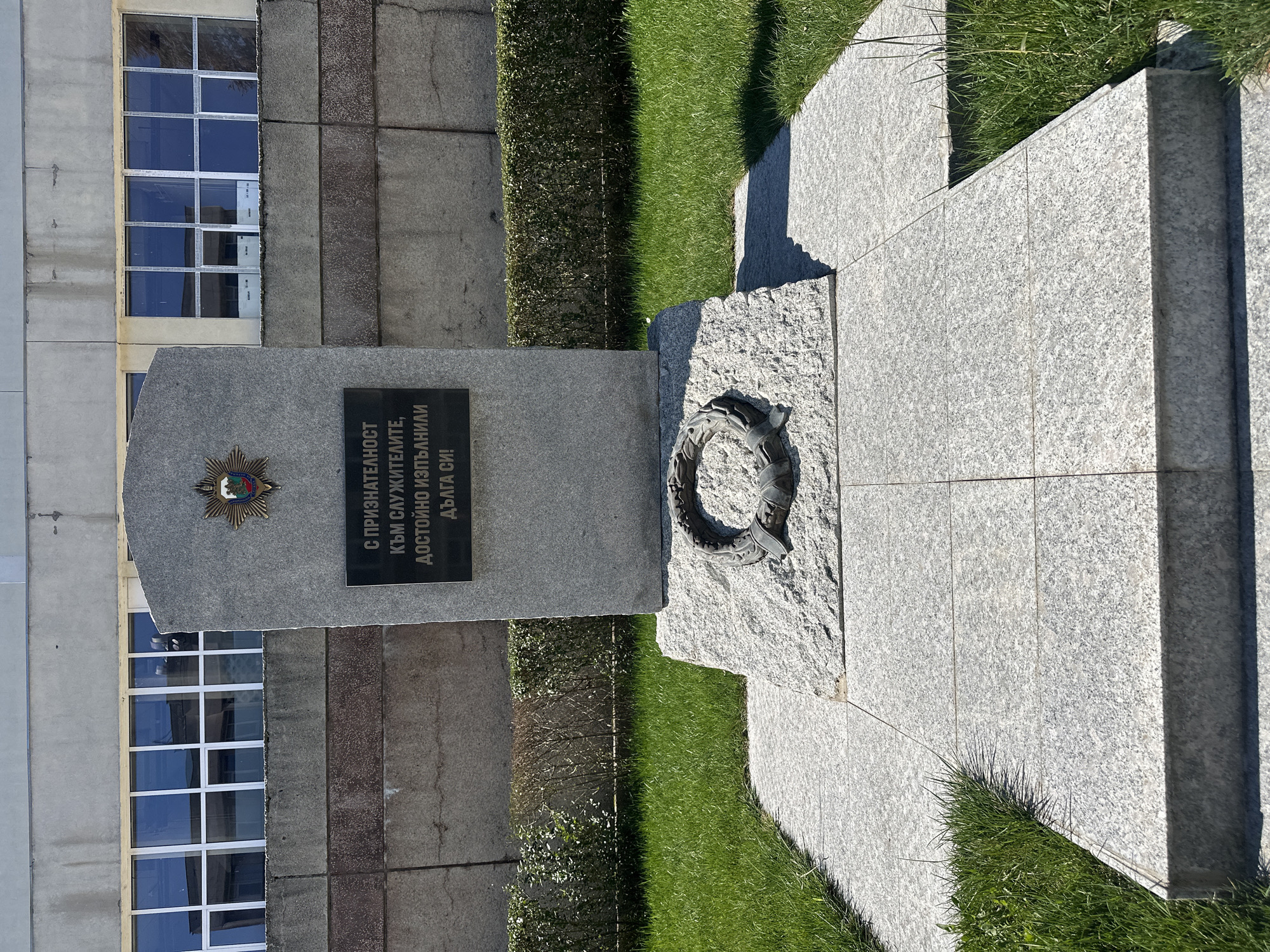 The image shows a stone monument located in Sofia, Bulgaria. The monument is composed of gray stone and features a plaque with Bulgarian text, an emblem, and a wreath. The text on the plaque translates to "With gratitude to those who served, performed their duty with dignity and died for it." The monument is surrounded by grass and a low hedge. Behind the monument is a building with multiple windows. The scene is bathed in sunlight, indicating it is daytime.