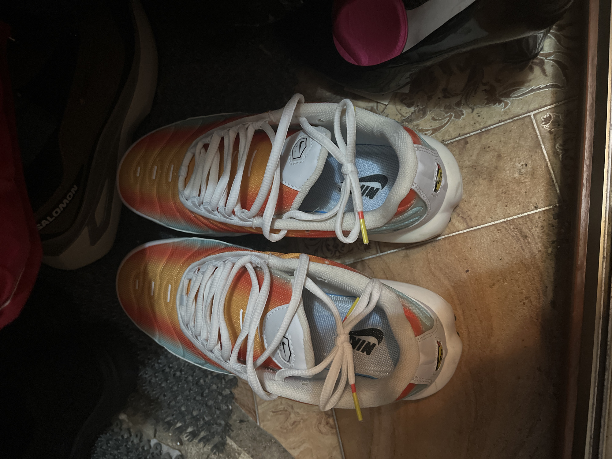The image shows a pair of Nike Air Max Plus sneakers with a gradient color scheme of orange, yellow, and teal on a tiled floor. The sneakers are tied with white laces, and a small yellow tag with red and yellow accents is visible on the laces of the left shoe. In the background, other footwear is partially visible, including a Salomon shoe. The scene appears to be indoors, possibly in a closet or storage area. There is no visible indication of the city of Minsk, Belarus.