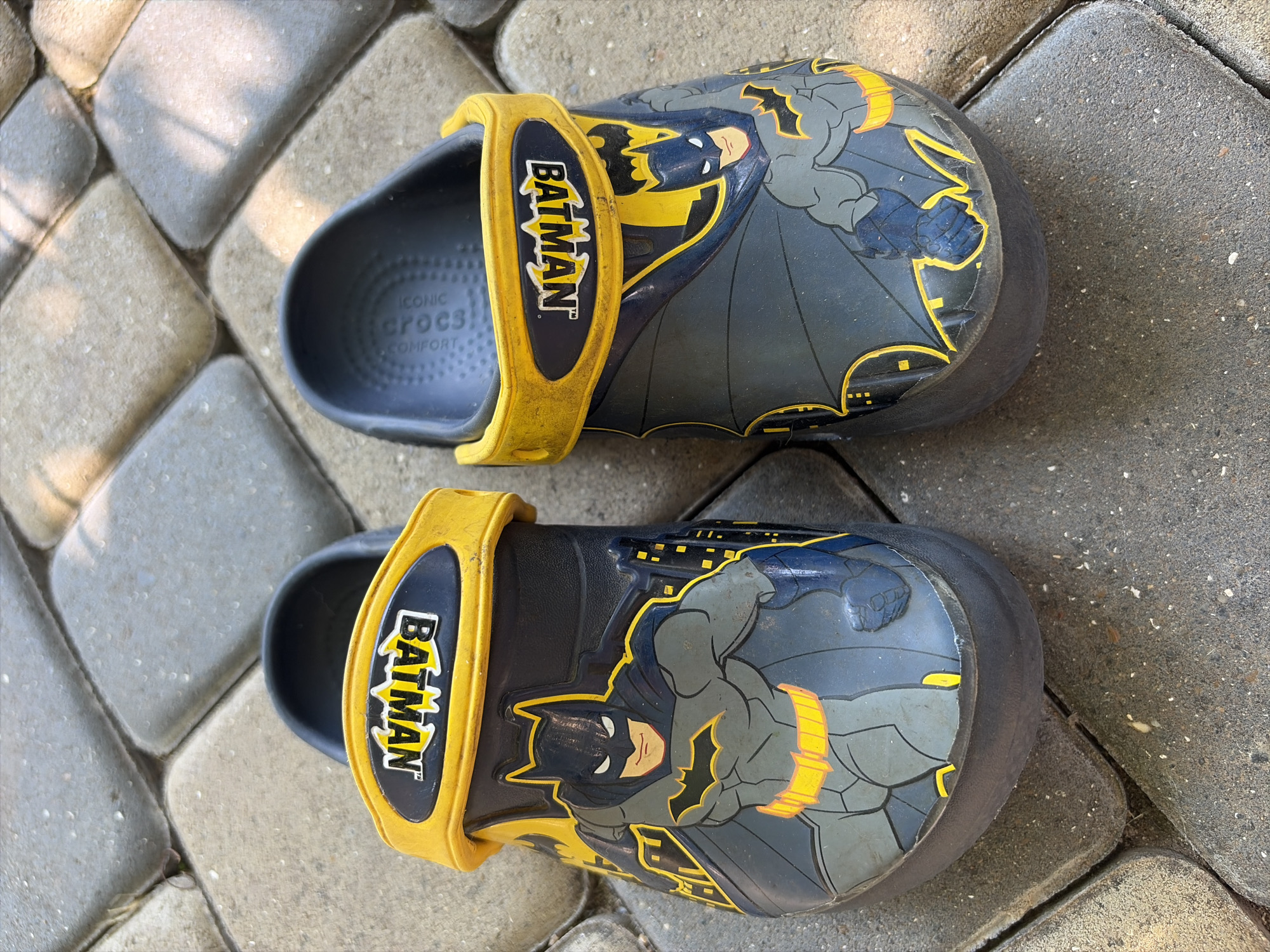 A pair of dark blue and grey Batman Crocs are on a stone-paved surface. The shoes have yellow straps with the word "BATMAN" in yellow letters within a black rectangle. The front of each shoe features an image of Batman in his suit. The scene is outdoors, with natural lighting. The location is Oxentea, Moldova.