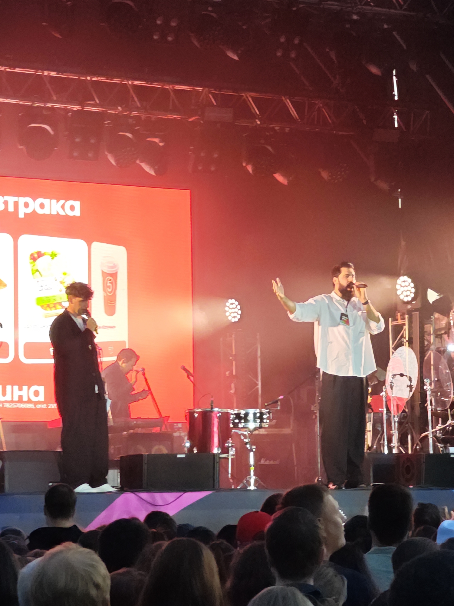On an outdoor stage in Yekaterinburg, Russia, a performance is underway. Two men are on stage, one in the foreground wearing a long black coat over a white shirt and dark pants, holding a microphone and speaking. The other man, to his left, is wearing a black robe and also holding a microphone. Behind them, a drum set is visible, and a large red screen displays graphics, one of which features a cup with the number "5". Bright stage lights illuminate the scene, casting a red glow. In front of the stage, a crowd of people watch the performance. The overall atmosphere appears to be that of a lively concert or event.