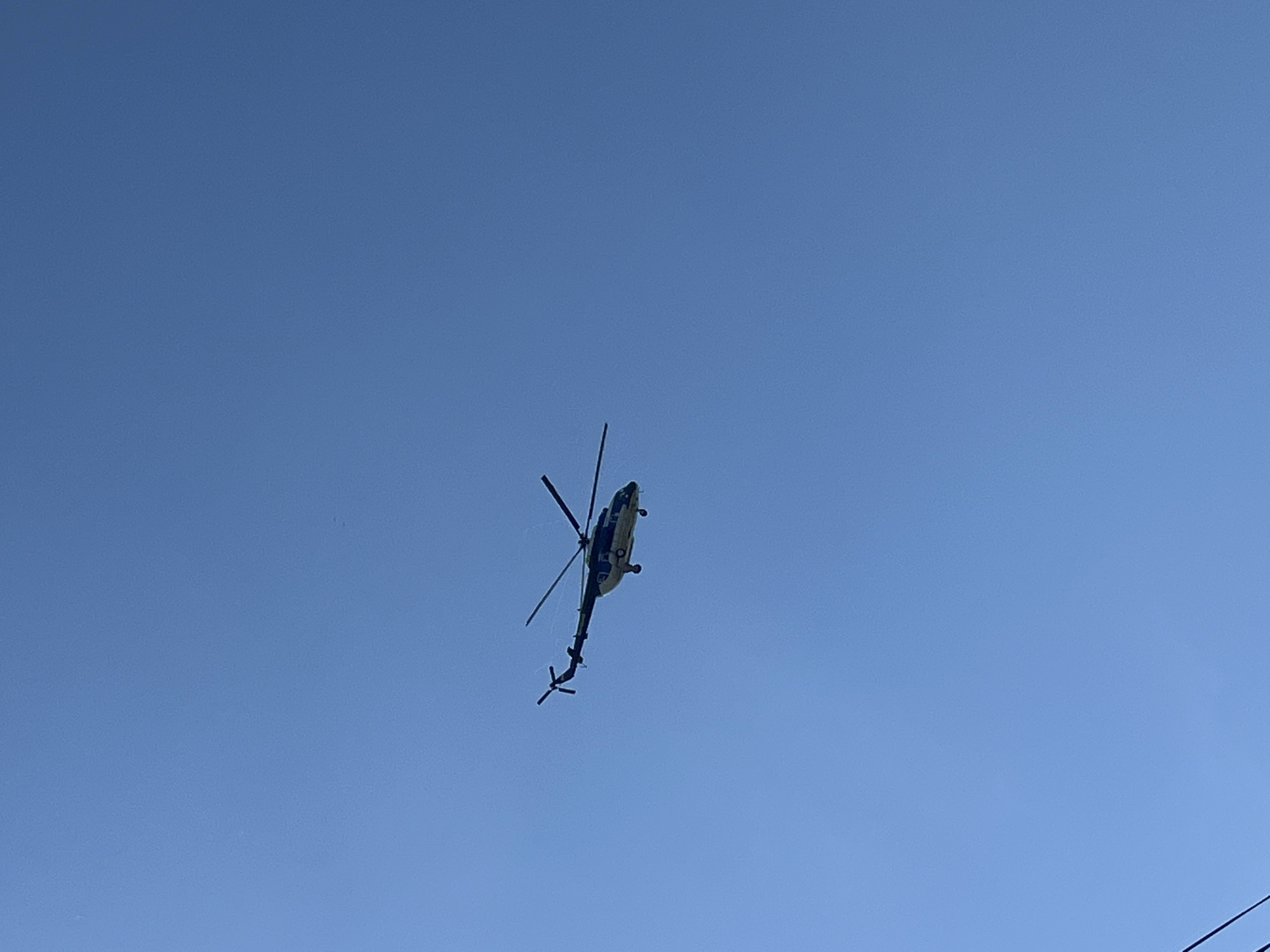 A helicopter is flying in a clear blue sky. It is difficult to determine the specific type of helicopter. The location is unknown and cannot be determined from the image alone, therefore information about the city of Chaikovsky, Russia cannot be confirmed.