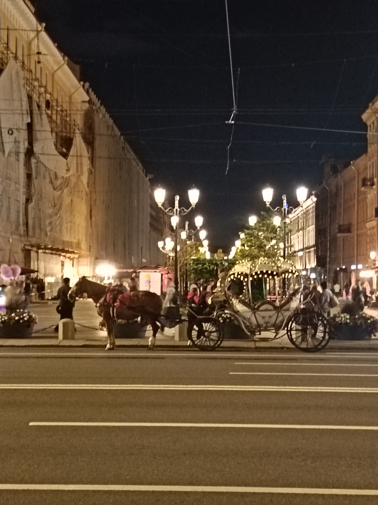 This image depicts a nighttime scene in Saint Petersburg, Russia, likely near Nevsky Prospekt, given the architectural style and the presence of horse-drawn carriages, which are a common tourist attraction in the city.
In the foreground, a horse-drawn carriage, possibly for tourism, is visible. The carriage itself is ornate, with a light-colored body and decorative lighting along its roof. It has large spoked wheels. The carriage is harnessed to a horse, which is brown and appears to be standing still. A person, likely the driver, is seated on the carriage.
Further to the left, another horse is hitched to a lighter, simpler carriage, with pink balloons attached to its front. A person in dark clothing stands near this carriage, holding the horse's reins.
The background features buildings with ornate facades, some of which appear to be under renovation, as indicated by scaffolding and draped coverings. Several streetlights, characteristic of Saint Petersburg's historic avenues, illuminate the scene. The sky is dark, indicating nighttime. The street in the foreground is paved and marked with white lines, suggesting it is a road for vehicular traffic. People are visible in the background, walking along the sidewalk and near the carriages. The overall atmosphere is lively and festive, typical of a tourist area at night.