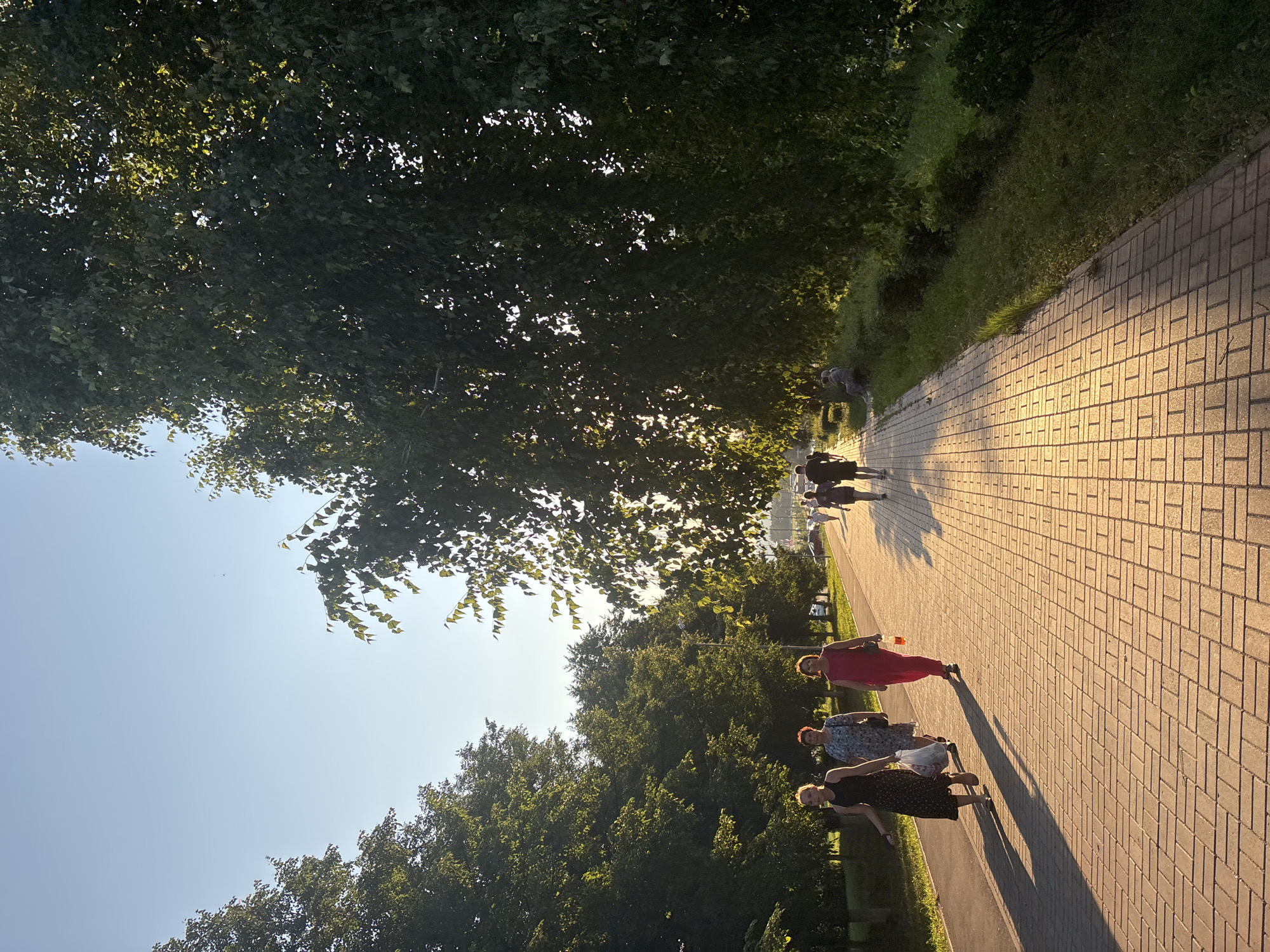 The image depicts a paved walkway in a park-like setting with trees lining the sides. The sun is low in the sky, casting long shadows and illuminating the brick-patterned walkway with a warm, golden light. Several people are walking along the path. On the left, three women are walking towards the camera. The woman closest to the viewer is wearing a dark patterned dress, the second woman is wearing a blue and white floral dress, and the third woman is wearing a bright pink jumpsuit and carrying a clear bottle with an orange liquid. Further down the path, a few more figures are visible, including some people sitting on a bench on the right side of the walkway. The trees are lush and green, suggesting a warm season. The sky is clear and blue. The scene appears to be a public area, possibly a city park, in Naberezhnye Chelny, Russia, given the presence of people enjoying an evening stroll in a well-maintained outdoor space.