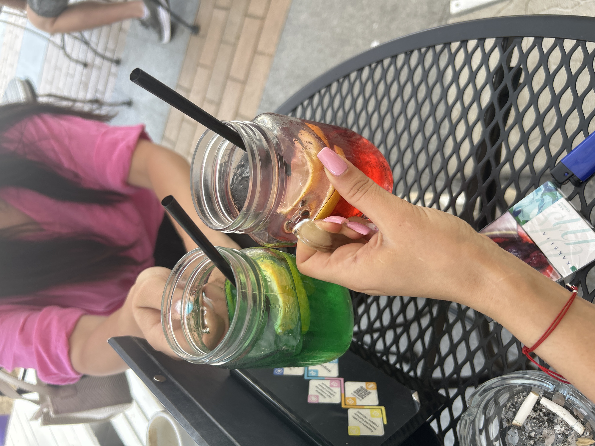 Two mason jar glasses, one filled with green liquid and a slice of lemon, the other with red liquid and slices of lemon, are held by unseen individuals. Both glasses contain black straws. The glasses are placed on a black metal mesh table. In the foreground, a hand with pink painted fingernails holds the glass of red liquid. A red string bracelet is worn on the wrist of another person whose arm is visible. An ashtray with cigarette butts, a pack of Karelia cigarettes, and a blue lighter are also on the table. In the background, a person in a pink shirt is partially visible, along with a tan tote bag. The scene appears to be outdoors, possibly on a patio. The ground is paved with bricks.