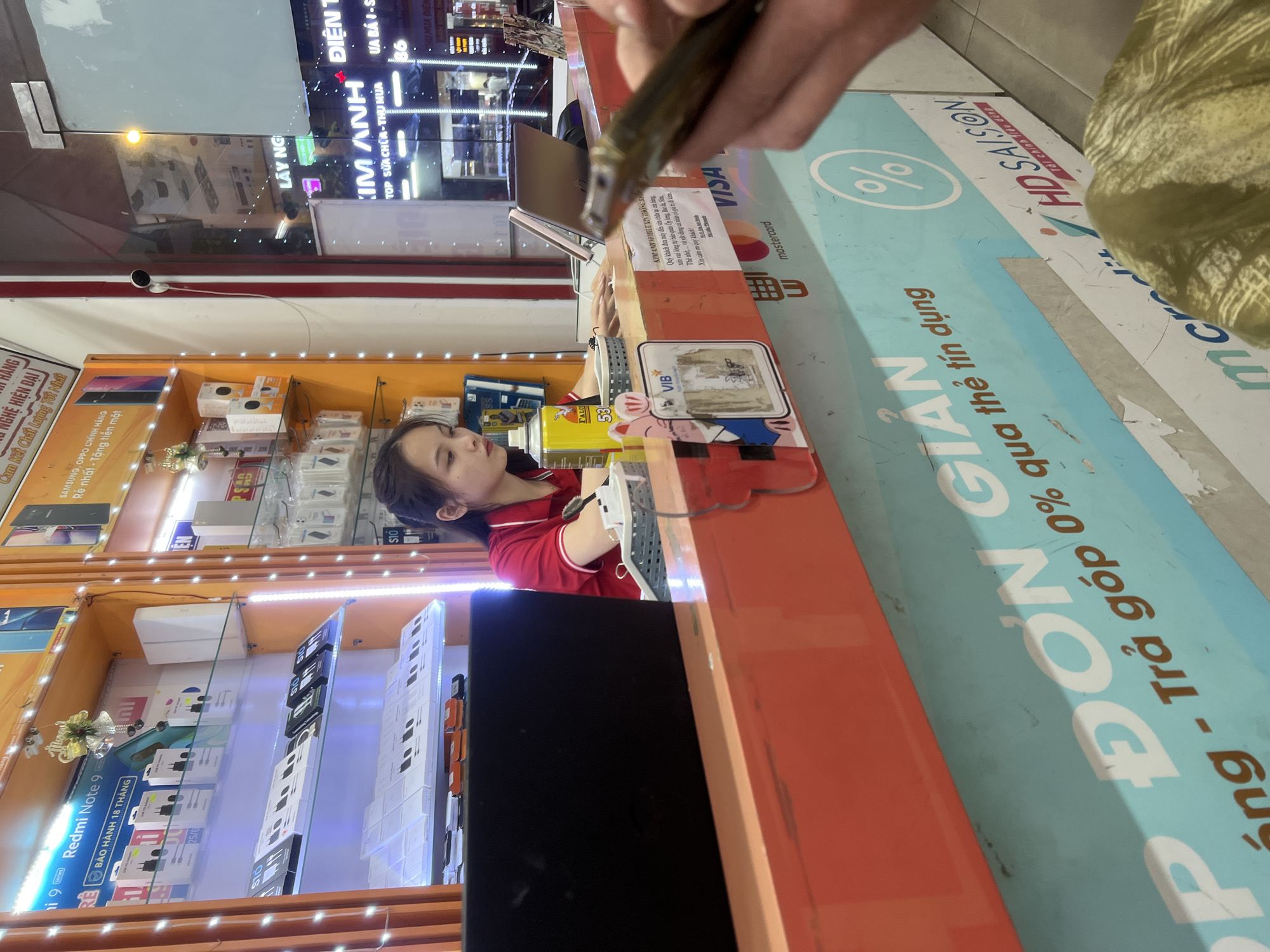 This media file captures a scene inside a mobile phone store in Da Nang, Vietnam. The primary subject is a young woman wearing a red polo shirt, likely an employee, standing behind a counter. She is looking down at a laptop or device on the counter, her expression calm and focused. In the foreground, a hand is holding up a mobile phone or similar electronic device, possibly to be examined or repaired.
The setting is a brightly lit retail environment with shelves stocked with various mobile phones, accessories, and display boxes. The shelves are illuminated by LED strip lights, creating a vibrant atmosphere. Visible products include Samsung and OPPO phones, as well as Redmi Note 9 accessories, highlighted by Vietnamese text such as "BẢO HÀNH 18 THÁNG" (18-month warranty) and "Rẻ nhất - Tặng tiền mặt" (Cheapest - Free cash gift).
Prominently displayed on the counter and along the wall are various signs and banners with text in Vietnamese and English. These include payment method indicators like "VISA" and "mastercard," along with branding for "HD SAISON" and "Tài chính 460 dùng" (Finance 460 uses), suggesting installment or financing options. Another large banner proclaims "ĐƠN GIẢN - Trả góp 0% qua thẻ tín dụng" (Simple - 0% installment via credit card). The name "KIM ANH MOBILE" is also visible, identifying the store. The overall impression is that of a busy, modern electronics store catering to customers with various purchasing and service needs. The time of day is not explicitly indicated, but the indoor lighting suggests it could be during business hours.