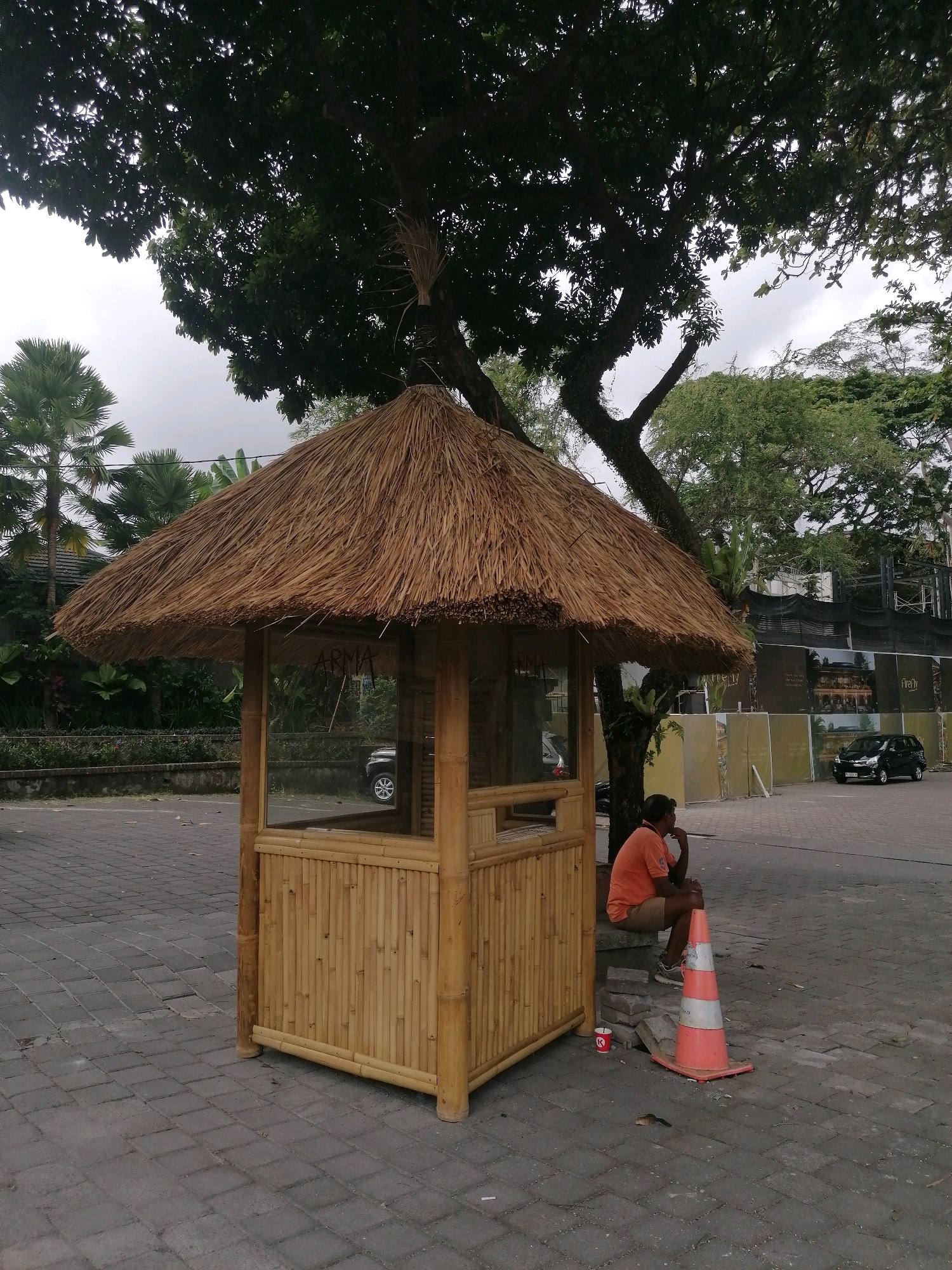 In the Ubud District, Indonesia, a person in an orange shirt and tan shorts is seated on a stone ledge next to a bamboo structure with a thatched roof. A traffic cone is positioned in front of the person. The bamboo structure appears to be a kiosk or booth with glass windows. In the background, a black SUV is visible. The ground is paved with interlocking gray bricks. Lush green trees and foliage are present, and a construction site with black netting and billboards is visible in the distance. The sky is overcast.