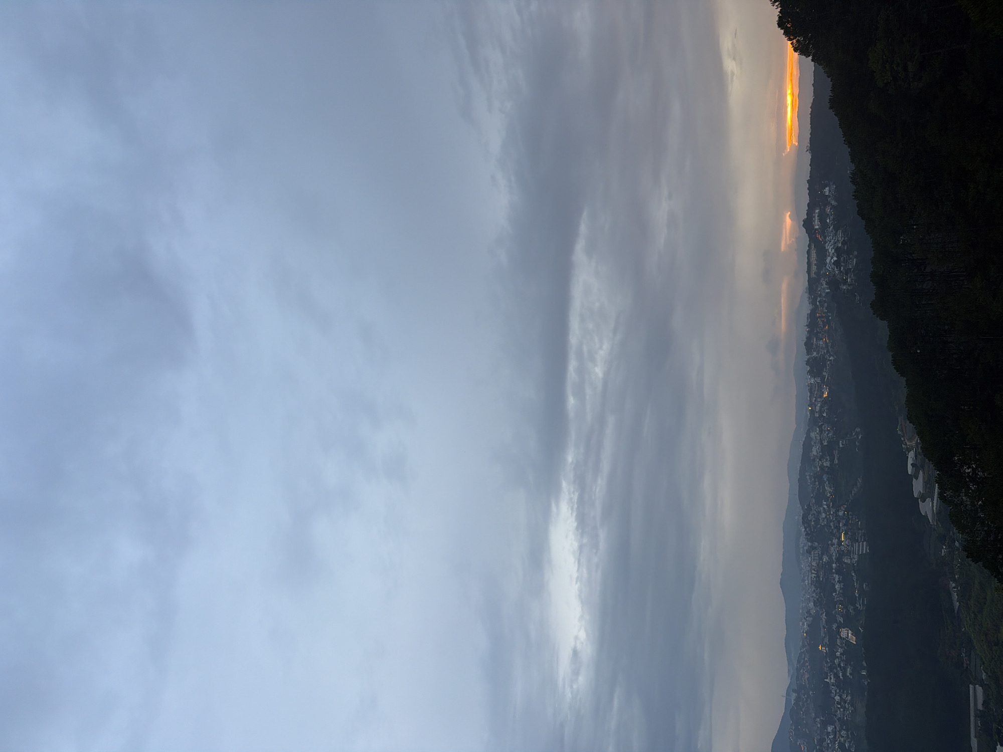 The image captures a panoramic view of a city nestled in a valley, likely Xuân Thành, Vietnam, at dusk. The sky is overcast with layers of gray clouds, with a sliver of vibrant orange light breaking through the horizon, indicating the setting sun. The cityscape below is densely populated with buildings of varying sizes and colors, many illuminated by artificial lights, creating a warm glow against the darkening landscape. The city is built on rolling hills, covered with lush green vegetation, and coniferous trees dominate the foreground, casting long shadows. The scene evokes a sense of tranquility and the quiet transition from day to night in a picturesque mountainous region.