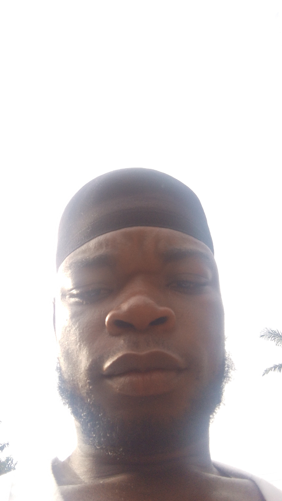 A close-up selfie shows a dark-skinned man wearing a dark-colored durag. He has a short beard. The background is heavily overexposed, obscuring environmental details, but some trees are vaguely visible in the lower right and left corners. The photo provides insufficient information to determine the precise location within Enugwu-Ukwu, Nigeria. No interactions or actions are discernible.