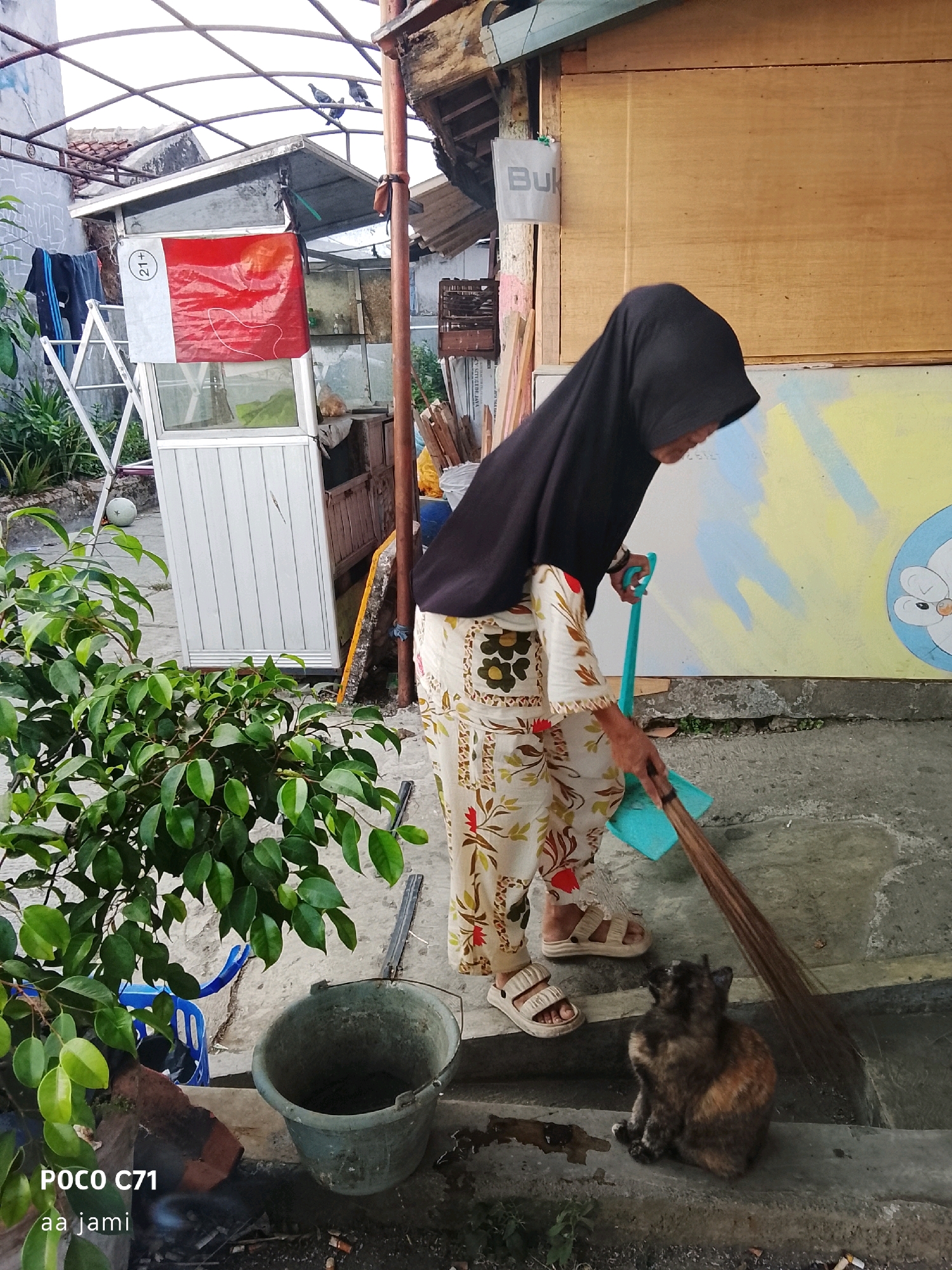 An elderly woman wearing a black hijab and a patterned dress is sweeping the ground with a broom. She is standing on a concrete surface, and a tortoiseshell cat is sitting nearby. In the background, there is a small white building with a red and white sign. The scene appears to be an outdoor area in Tasikmalaya, Indonesia, possibly a street or a yard, with some vegetation and debris visible. The overall setting suggests a simple, everyday scene.