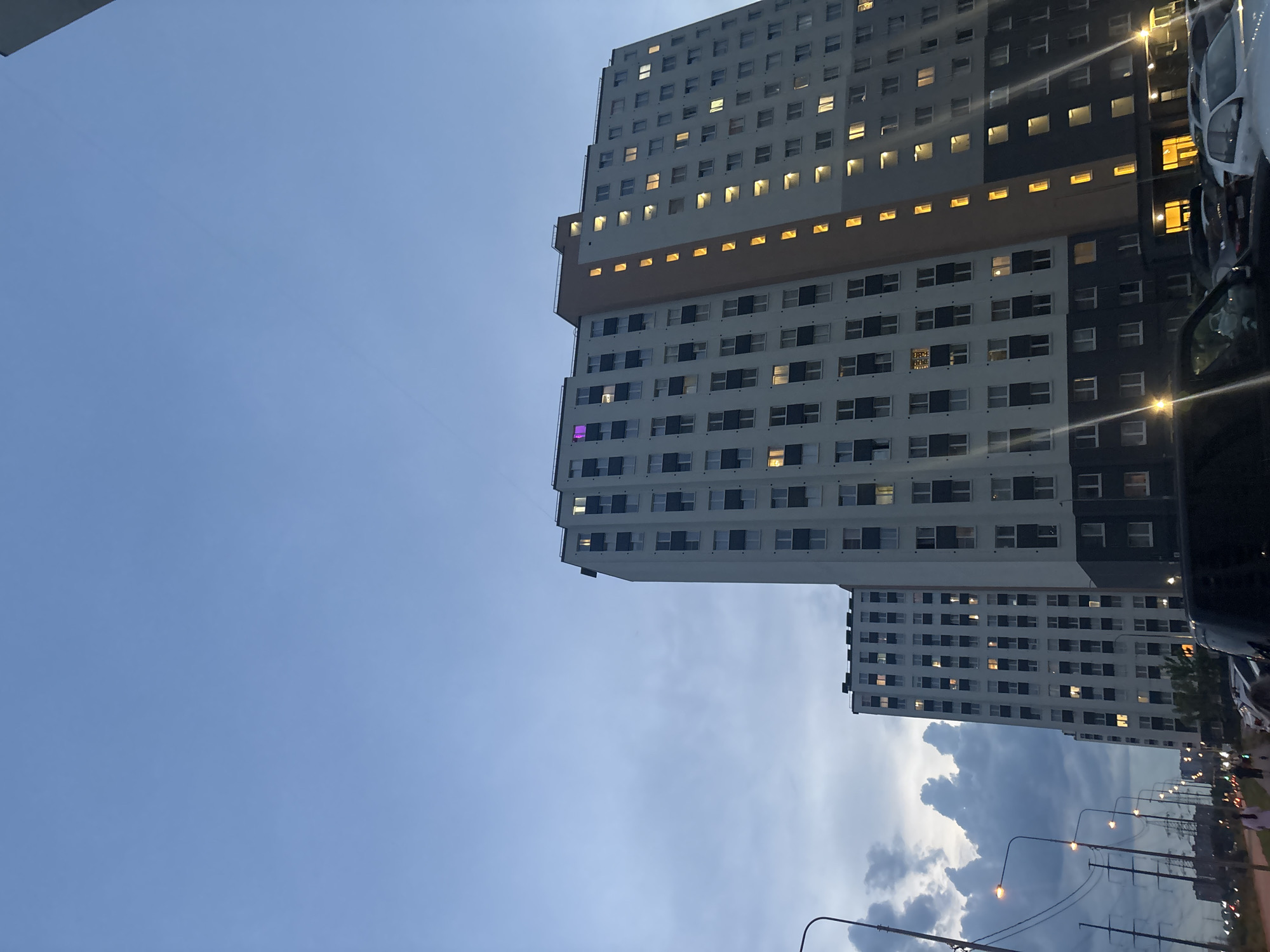The image depicts an urban twilight scene in the city of Unknown, Russia, centered on two multi-story residential buildings. The prominent building on the right features a white and dark grey facade with a vertical orange-brown architectural element; approximately 15 floors are visible. Numerous windows exhibit warm yellow interior illumination, with one upper-story window displaying a distinct purple light. A second, similar residential building stands behind and to the left. The sky above is overcast, presenting a blue-grey hue with some textured clouds, particularly on the left side of the frame. In the mid-ground and foreground on the left, a street is illuminated by multiple streetlights emitting yellow light, alongside utility poles and overhead power lines. Indistinct human figures are present on a paved walkway beside the street. Several vehicles are parked on the street and in the immediate foreground, including a dark-colored SUV partially obstructing the lower right portion of the image.