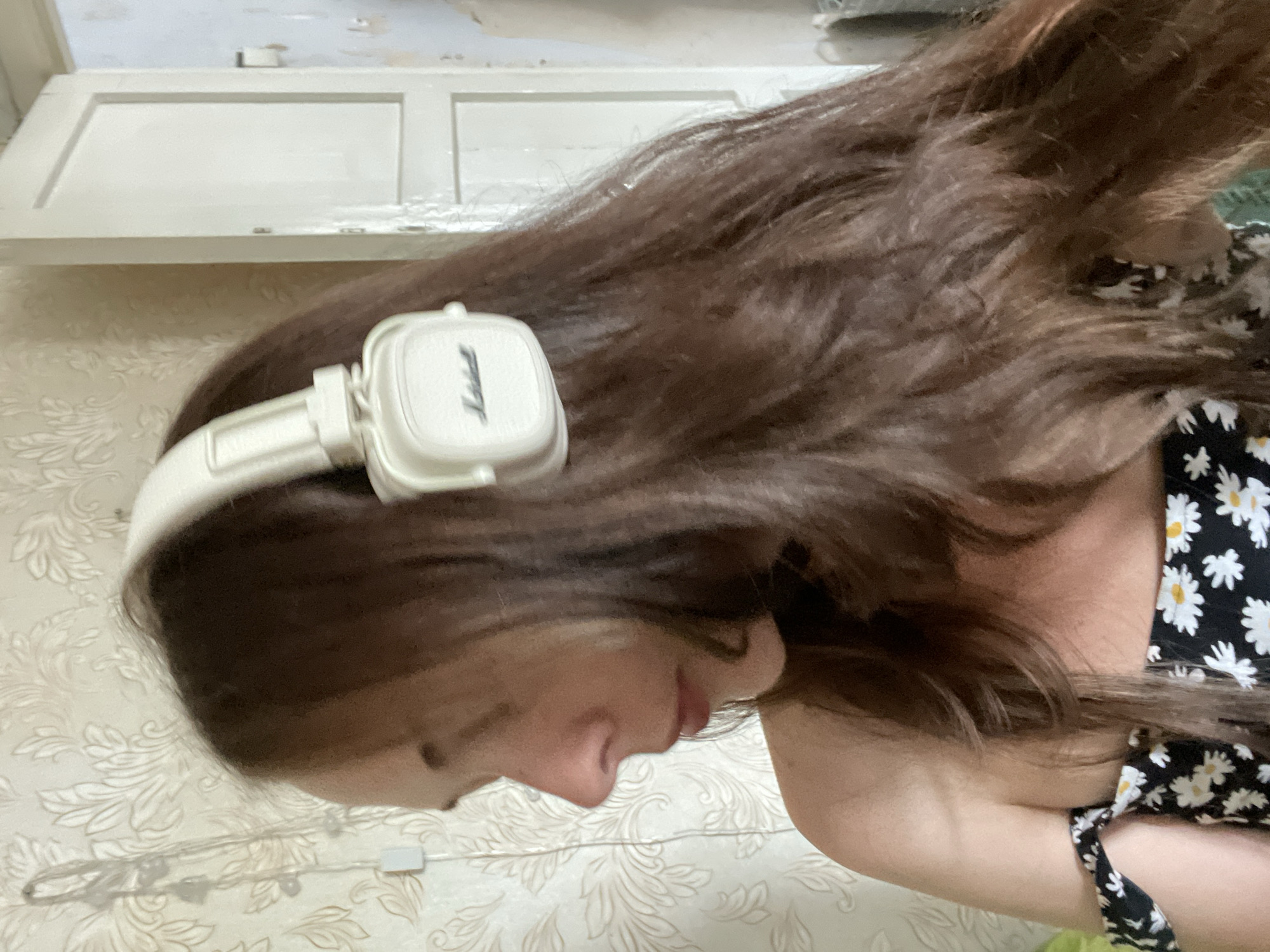 A medium shot shows a woman from the shoulders up, wearing white Marshall headphones. She is facing left, with her head tilted down. Her long, dark brown hair is visible. She is wearing a black and white floral print off-the-shoulder top. The background consists of patterned wallpaper and a white door. The lighting is dim. The location is unknown.