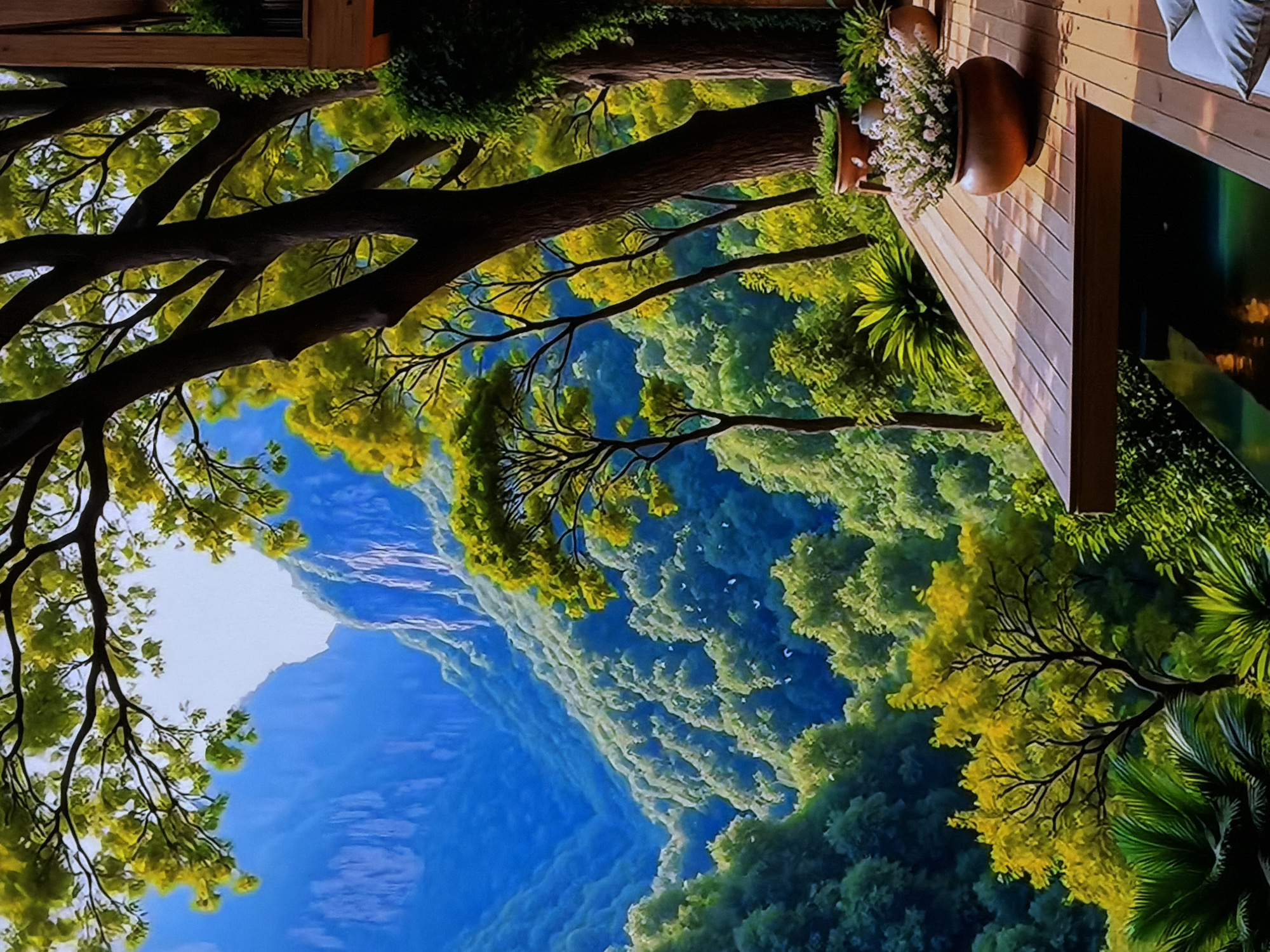 The image displays a vibrant, serene mountainous landscape, viewed from a high vantage point, likely a wooden deck or balcony. In the immediate foreground, a warm-toned wooden deck is visible, featuring several potted plants. One large terracotta-colored pot holds delicate white or light purple flowers, while others contain lush green foliage, including a palm-like plant. A white cushion or pillow hints at outdoor seating. A dark, reflective surface at the edge of the deck might be a small pool or a screen.
Beyond the deck, a dense forest unfolds with large trees, their dark trunks and bright green to yellowish-green foliage filling the midground. The branches of these trees frame a panoramic view of rolling forested hills and mountains receding into the distance. The sky above is a clear, bright blue, with a stark white area suggesting intense sunlight.
No people are visible, and no specific activity is taking place, creating a peaceful and contemplative atmosphere. The scene is set during daytime under clear, sunny weather. The vivid colors and slightly stylized textures, particularly in the distant landscape, suggest the image could be a digital rendering, a large-scale artistic mural, or a high-resolution display, rather than a direct photograph of a natural outdoor scene. There is no visible text in the image. The location context provided is Sofiivska Borshchahivka, Ukraine.