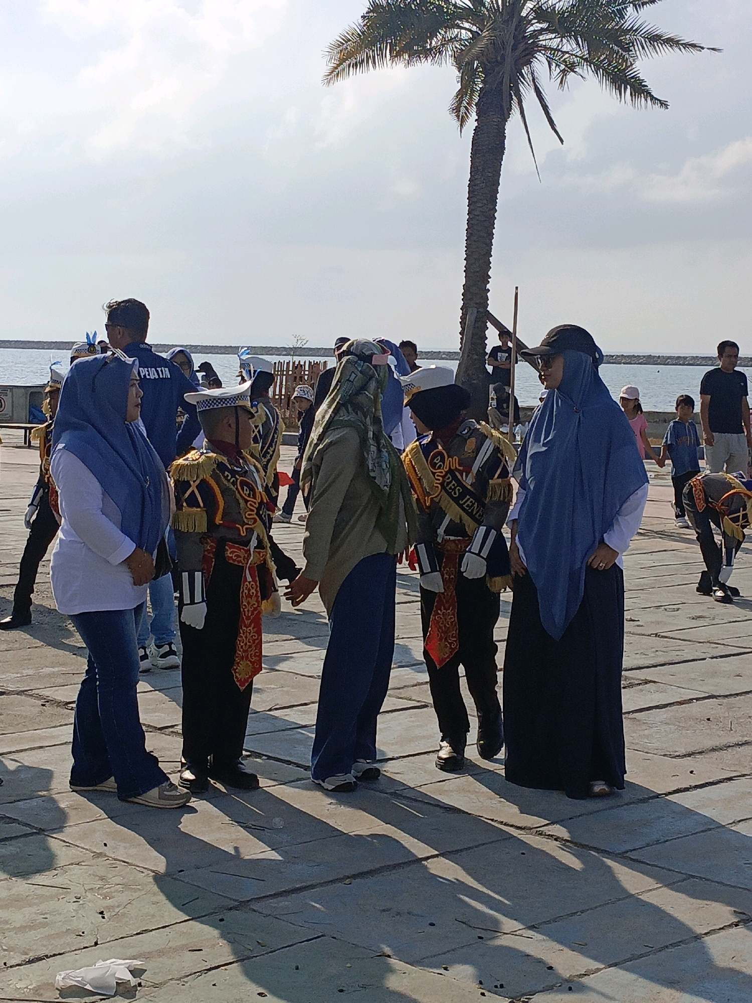 The image depicts an outdoor scene on a paved area near a body of water, likely the sea, under a bright sky with scattered clouds and a prominent palm tree. The location is identified as Bulukumba, Indonesia. Several individuals are present, many of whom are dressed in formal or ceremonial attire.
In the foreground, a woman wearing a blue hijab and a white long-sleeved shirt is standing and looking towards her right. Next to her, a person in a formal uniform, possibly a military or ceremonial cadet uniform with a white cap, gold epaulets, and a red and gold sash, is facing to the right. Another person in a tan headscarf and green jacket is facing the person in uniform. Further to the right, a woman in a dark blue hijab and a dark skirt stands, also looking towards the right.
In the background, several other individuals are visible, including adults and children. Some are wearing dark t-shirts, and some children are in casual clothing. There are structures in the background that appear to be part of a waterfront area, possibly a pier or boardwalk, with railings. The ground is a paved surface made of rectangular tiles. Shadows from the people and the palm tree are cast on the ground, indicating strong sunlight. A crumpled white paper object is on the ground in the lower left corner. The overall impression is of a public gathering or event taking place outdoors.