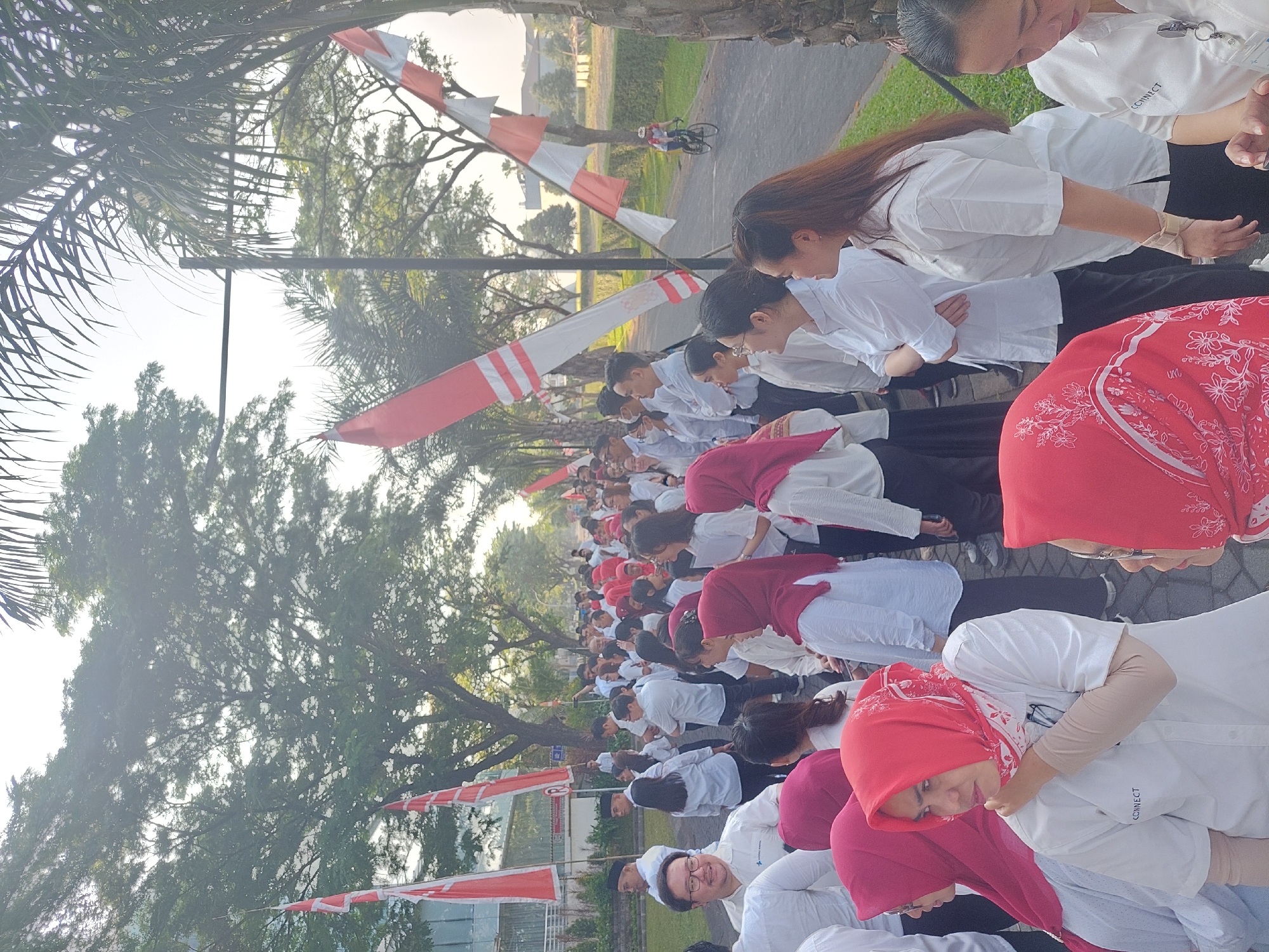 In a city street scene in Surabaya, Indonesia, a large group of people, predominantly dressed in white shirts and some in red hijabs, are gathered outdoors. The setting appears to be a pathway lined with trees and flags, suggesting a formal event or celebration. Red and white striped flags are prominently displayed on poles along the street, consistent with the Indonesian national flag. In the background, buildings and more trees are visible, indicating an urban or semi-urban environment. Several individuals are seen interacting, talking, and looking in various directions, while others stand in organized lines. The overall atmosphere seems festive and communal, with a sense of shared activity. A person on a bicycle is seen in the far background, riding on the adjacent road. The lighting suggests it is daytime, likely morning or late afternoon given the angle of the sun.
