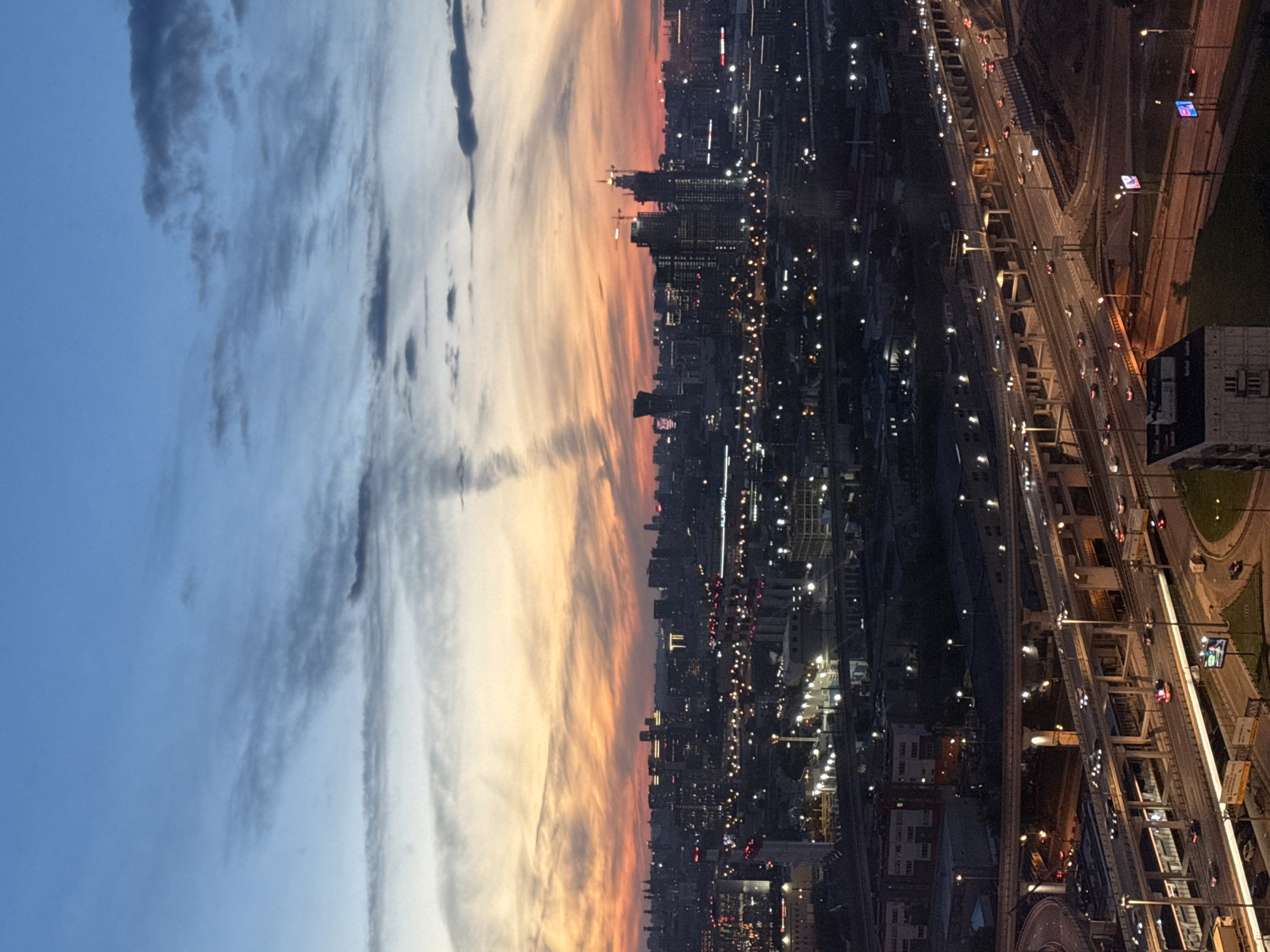 An aerial, vertical, medium shot captures a city skyline at dusk. The sky transitions from a deep blue at the top to hues of orange and pink near the horizon, adorned with wispy clouds. Below, a sprawling cityscape is illuminated by the warm glow of streetlights and building windows. A complex network of multi-level highways dominates the foreground, with vehicles visible on the roadways. Industrial buildings and residential structures are interspersed throughout the urban landscape. Construction cranes are present among some of the taller buildings in the mid-ground. The scene is devoid of any visible people or discernible interactions.