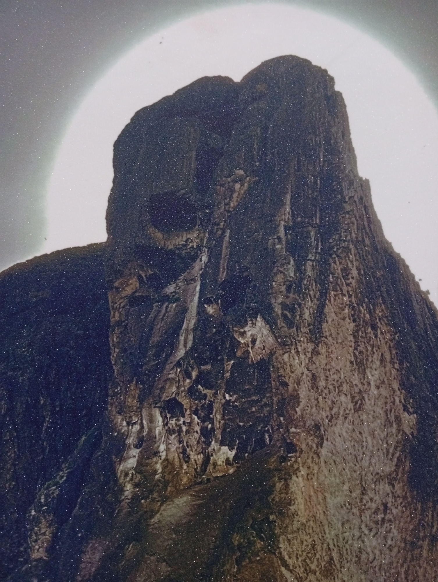 The image shows a rock formation that resembles a face. The rock is dark and appears to be made of rough, textured stone. It is set against a bright, hazy sky. The lighting suggests it may be early morning or late evening, or that there is a strong light source behind the formation, possibly the sun or moon, creating a halo effect. The formation is tall and imposing, with various crevices and shadows that contribute to the facial illusion. The image is grainy, suggesting it was taken with a low-resolution camera or under poor lighting conditions. The location is identified as Gura Top, Nigeria, a geographical feature known for its distinctive rock formations. There are no people or animals visible in the image.