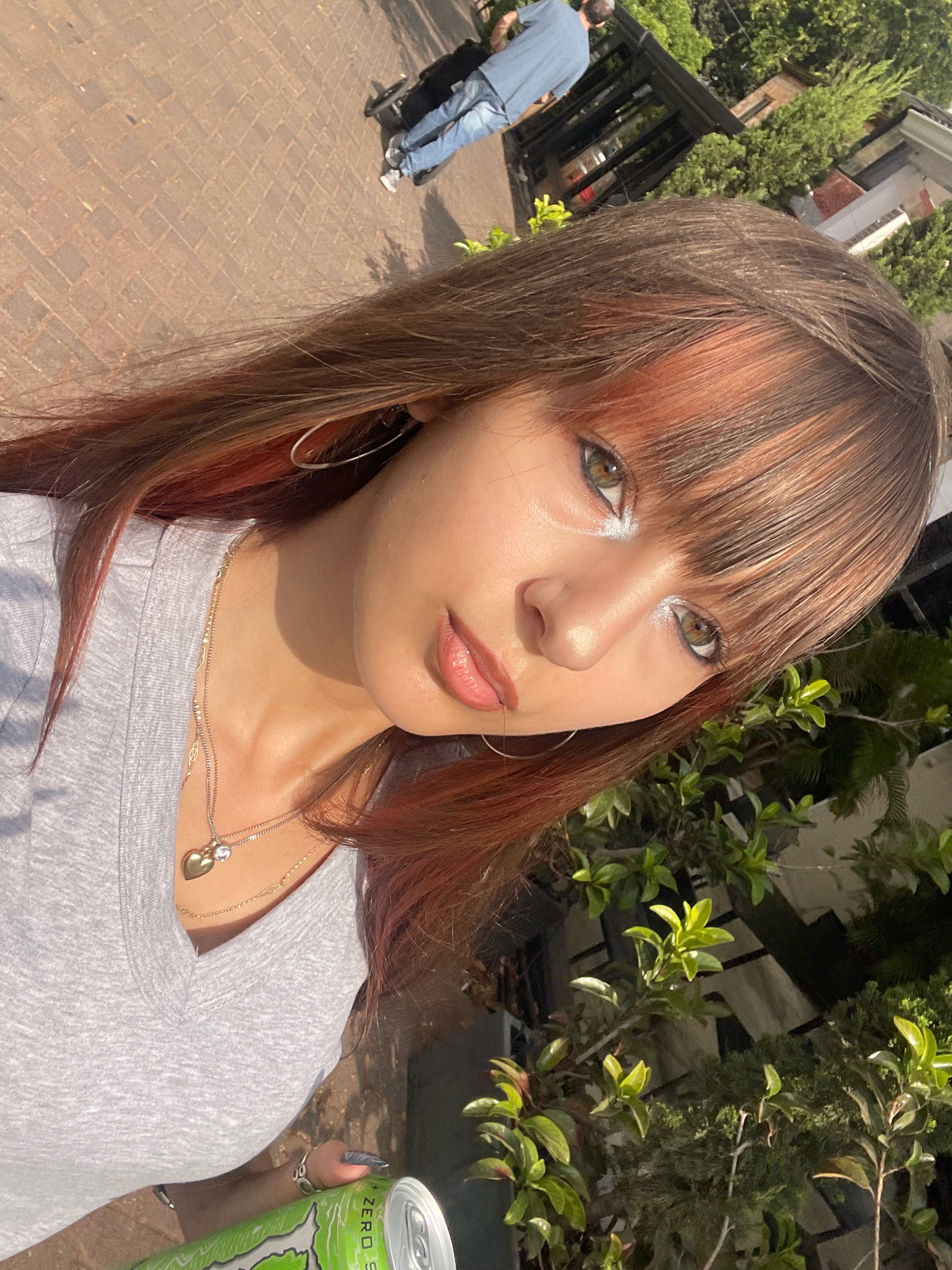 A young woman with reddish-brown hair and bangs is taking a selfie outdoors. She is wearing a gray t-shirt and several necklaces, one of which has a gold heart pendant. Her makeup includes white eyeliner. In her right hand, she holds a green can of Monster Energy drink. Behind her, a person in jeans and a blue shirt is visible, pushing a stroller or cart along a brick pathway. Lush green foliage and a building with dark accents are also present in the background. The lighting suggests it is daytime with strong sunlight. The setting appears to be a residential area or public space in Raanana, Israel, possibly a park or sidewalk.