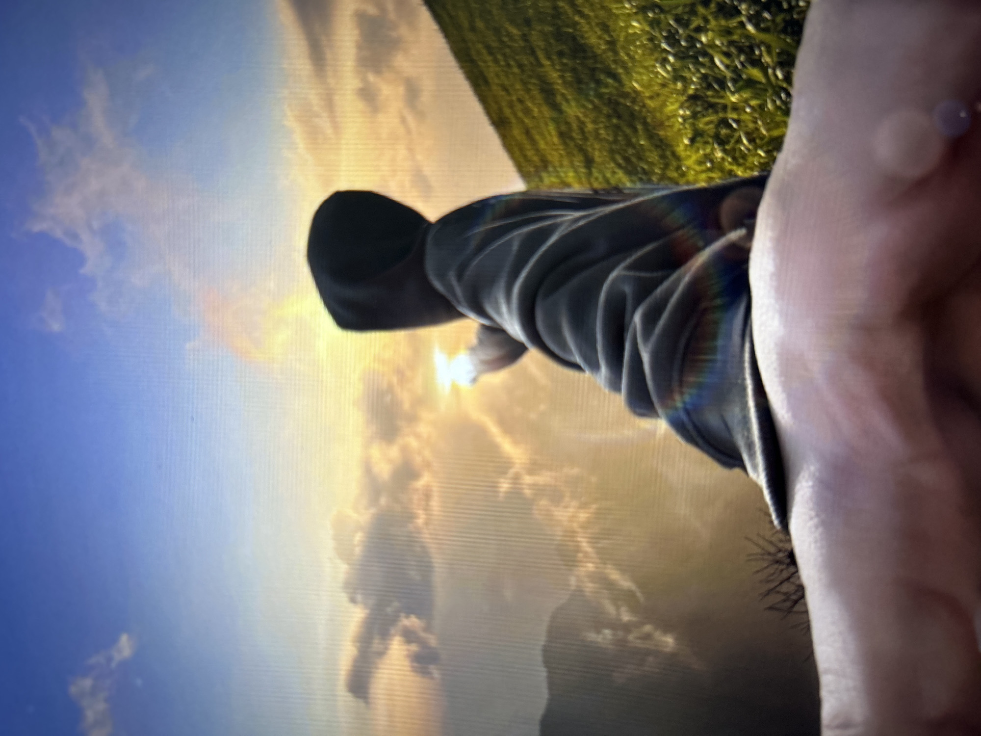 The image depicts a digital screen displaying a scenic landscape during either sunrise or sunset, specifically in the context of the city of Burgas, Bulgaria. One individual, seen from behind and slightly to the right, occupies the middle ground. The person wears a dark, hooded garment with the hood up, obscuring their head and face. Their right arm is extended towards a bright, intense light source, likely the sun, positioned towards the upper right of the frame, causing a strong glare and a rainbow-colored lens flare effect across their arm. The background features a vast sky transitioning from blue at the top to warm golden and orange hues near the horizon, populated by scattered clouds. Mountains or large hills are visible in the mid-ground on the left, partially obscured by atmospheric haze and clouds. On the right, a steep, vibrant green slope covered in dense vegetation descends. In the immediate foreground, occupying the bottom edge of the frame, is a blurry, light-skinned hand, likely belonging to the person photographing the screen. The visible pixel grid across the entire image confirms it is a photograph of a digital display.