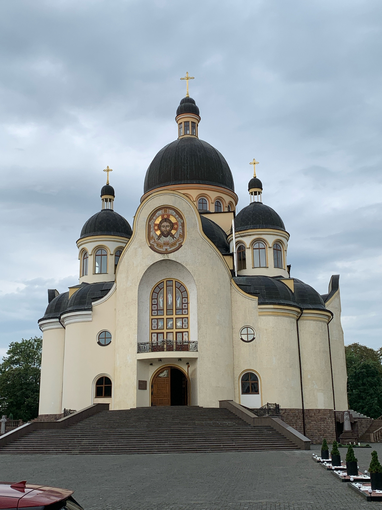 The photograph shows the Church of the Blessed Virgin Mary in Kolomyia, Ukraine. The church is a large, cream-colored building with multiple dark domes topped with golden crosses. The central dome is the largest, with two smaller domes on either side and two even smaller domes further out. The facade features a large arched entrance with wooden doors, above which is an arched window with stained glass depicting religious figures. A mosaic of Jesus is positioned above the stained-glass window. A balcony with a decorative railing is situated in front of the entrance. Wide stone stairs lead up to the church entrance from a paved courtyard. The sky is overcast, and trees are visible to the left and right of the building. Several potted evergreen trees line the paved area in the foreground. A red car is partially visible in the lower left corner of the image.