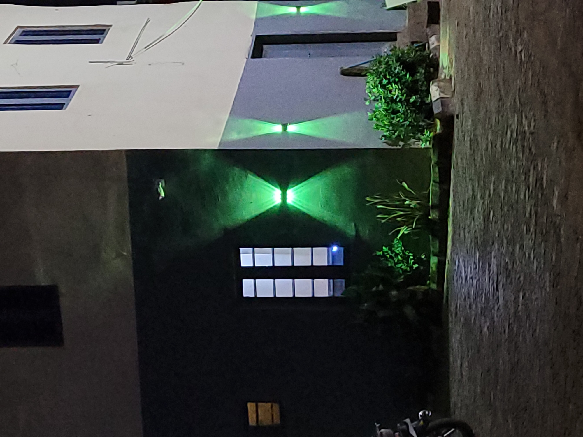 At night in Akwanga, Nigeria, a building with a white upper portion and a darker lower portion is lit by green lights. The building has rectangular windows, some illuminated and some dark. Green lights are mounted on the exterior, casting an "X" pattern on the walls. Plants grow near the building. A section of a paved walkway or road is visible on the right side of the frame, with the bottom of a vehicle partly visible at the bottom center.