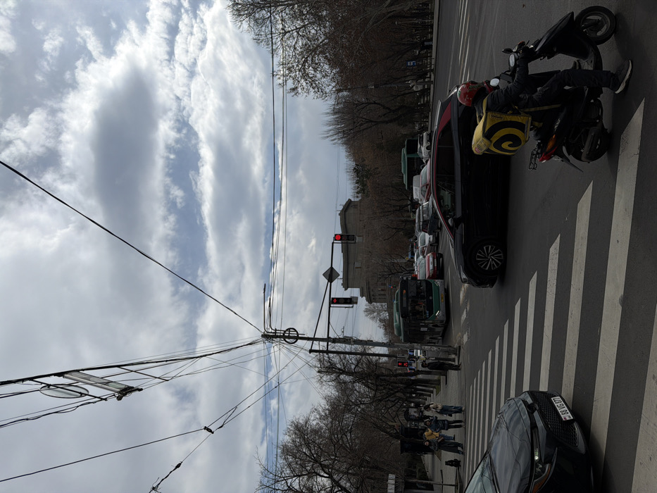 The image captures a street intersection on Baghramyan Avenue in Yerevan, Armenia, with the National Assembly of Armenia building visible in the background. The sky is largely overcast with areas of visible blue.
Overhead, a dense entanglement of black electrical and communication wires is strung between poles, with a street light also visible.
In the immediate foreground, a dark-colored Toyota Corolla with Armenian license plate "88 VG 077" is stopped at a white-striped pedestrian crosswalk on the left. To its right, a person wearing a dark jacket, dark trousers, and a red helmet sits on a black scooter, also stopped at the crosswalk. A large, square, bright yellow delivery bag with a prominent black spiral logo is worn on the individual's back.
Traffic at the intersection is halted by two visible red traffic lights. A dark SUV, featuring a subtle red trim line, is stopped in the right lane behind the scooter. In the adjacent left lane, a long green city bus, carrying passengers, is also stopped. Multiple other cars and a second green bus are visible in stationary traffic further down the road.
On the left sidewalk, a group of approximately five to six pedestrians stands near the crosswalk. One individual further to the left appears to be in a crouching or seated posture. The street is lined on both sides with bare deciduous trees, suggesting late winter or early spring. The prominent building in the distant background, partially obscured by trees and traffic, is a large, grey, multi-story structure with distinct architectural features, identified as the National Assembly of Armenia.