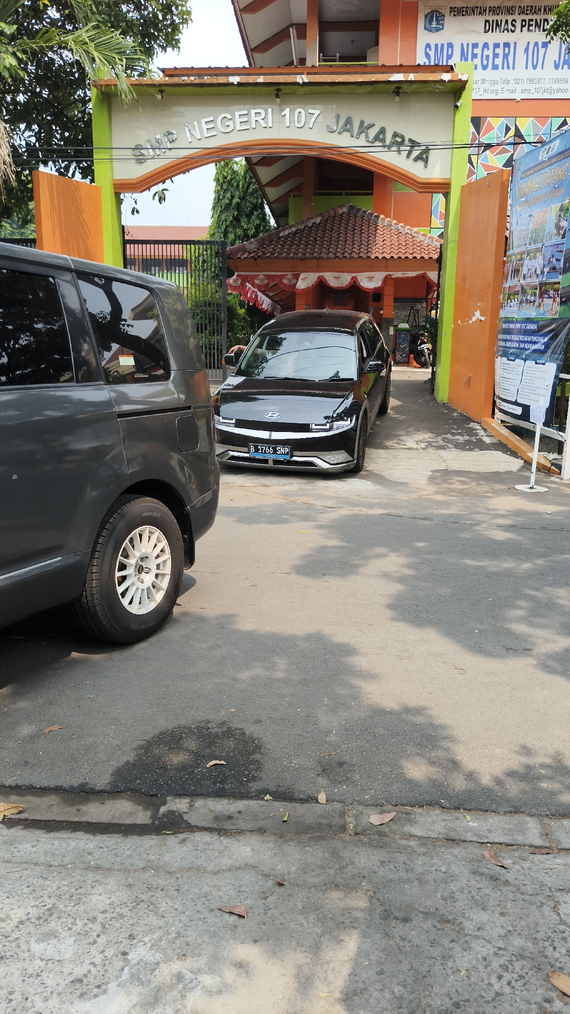 The image depicts the entrance of SMP Negeri 107 Jakarta, a public junior high school located in South Jakarta, Indonesia. The school's name is prominently displayed on an archway above the entrance. Two vehicles are visible: a dark grey minivan on the left and a black Hyundai Ioniq 5 on the right, parked in front of the school. The Hyundai Ioniq 5 has a license plate that reads "B 1766 SNP". The school building has orange and green architectural elements, and a welcome banner is displayed on the right side of the entrance. The ground is paved with asphalt, and there are some fallen leaves. The scene appears to be captured during daylight.