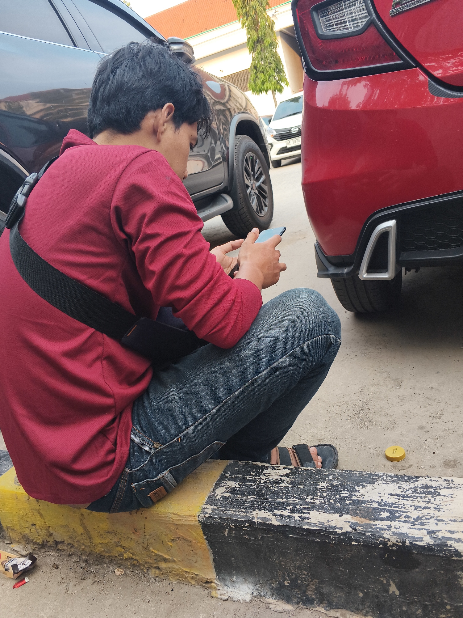 A young man in a red long-sleeved shirt and blue jeans is sitting on a curb in Bandar Lampung, Indonesia, looking at his phone. He is wearing black sandals and has a black strap across his chest. A red car is parked behind him, and a dark-colored SUV is to his left. The curb is painted yellow and black and appears worn. A small yellow cap and a crumpled cigarette pack are on the ground near the curb. The scene suggests an outdoor urban environment, likely a parking area or street side.