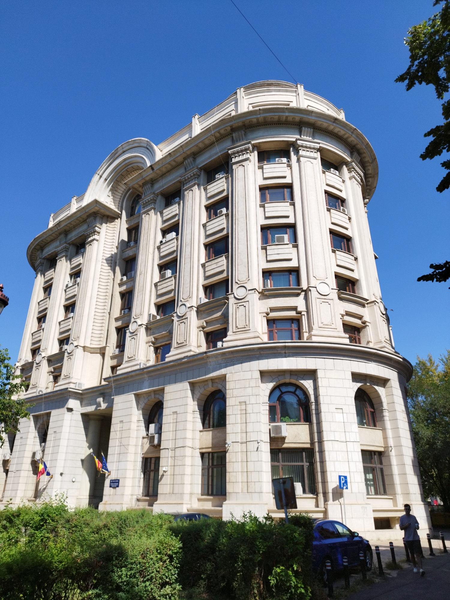 The image shows a large, light-colored, ornate building with a curved facade and a domed roof. The building has many windows and architectural details, including columns and decorative moldings. Two flags are visible on the lower part of the building, one Romanian flag and one European Union flag. A blue car is parked on the street in front of the building, partially obscured by bushes. A person is walking on the sidewalk near the car. The sky is clear and blue, indicating a sunny day. The building appears to be located in a city, likely Bucharest, Romania, given the presence of the Romanian flag.