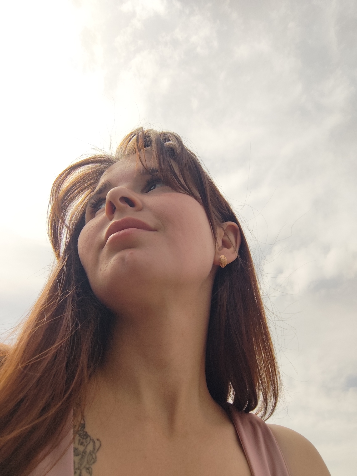 A low-angle shot shows a young woman looking upwards towards a cloudy sky. She has reddish-brown hair with bangs and is wearing a pink top. A small tattoo is visible on her chest. She is wearing stud earrings. The location is not discernible from the image.