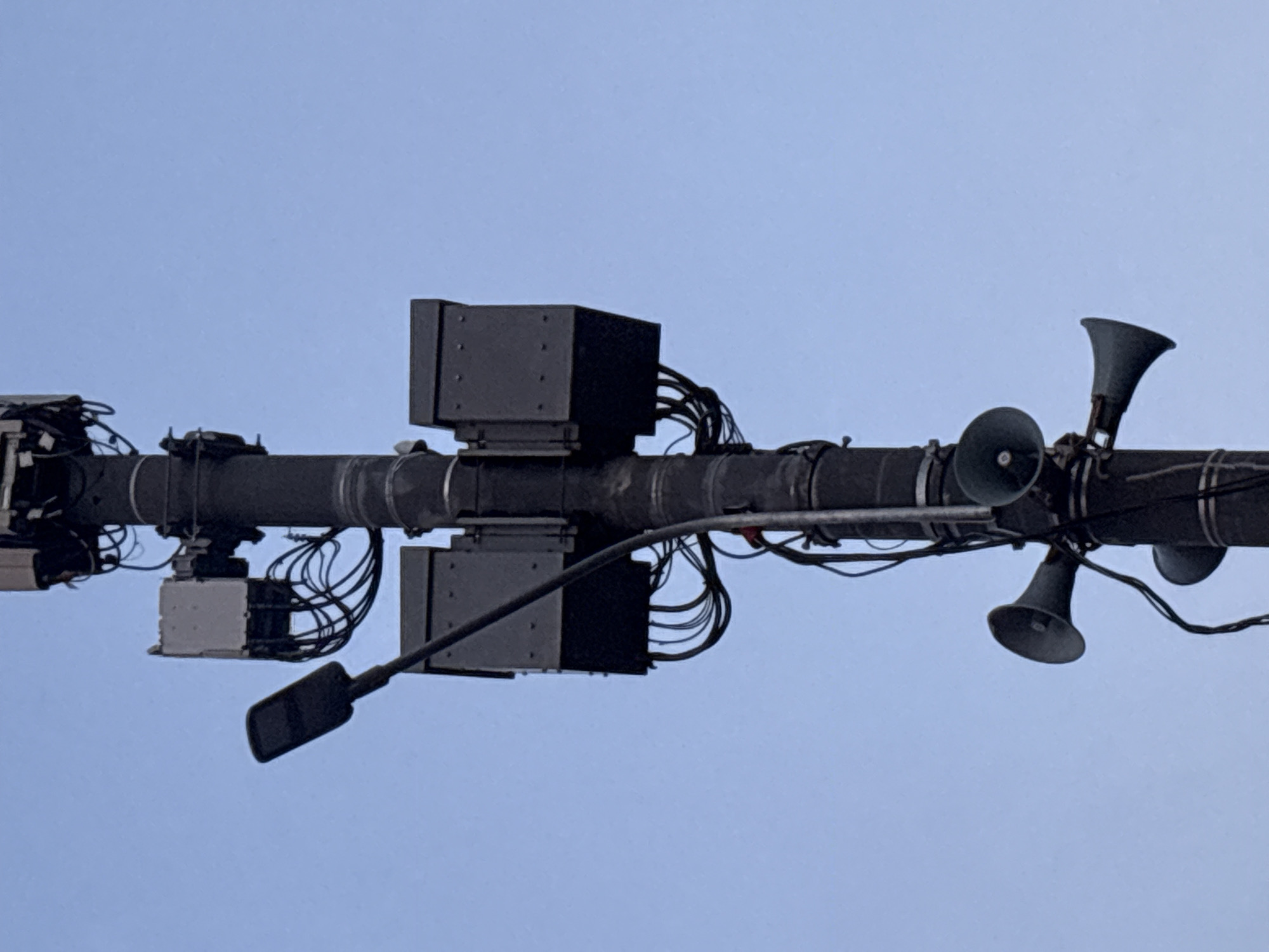 This media file is a close-up, eye-level shot of a dark, horizontal pole mounted against a clear, pale blue sky, suggesting it is daytime. Attached to the pole are various pieces of equipment. On the left side, there are several dark boxes and what appears to be a light fixture hanging down. Moving towards the center and right, there are more dark rectangular boxes with wires connecting them, and several dark, horn-shaped speakers pointed in different directions. The overall scene depicts an urban or industrial setting, possibly a streetlamp or utility pole with integrated communication or surveillance technology. There is no visible text in the image, and no people are present. The weather appears clear and calm.