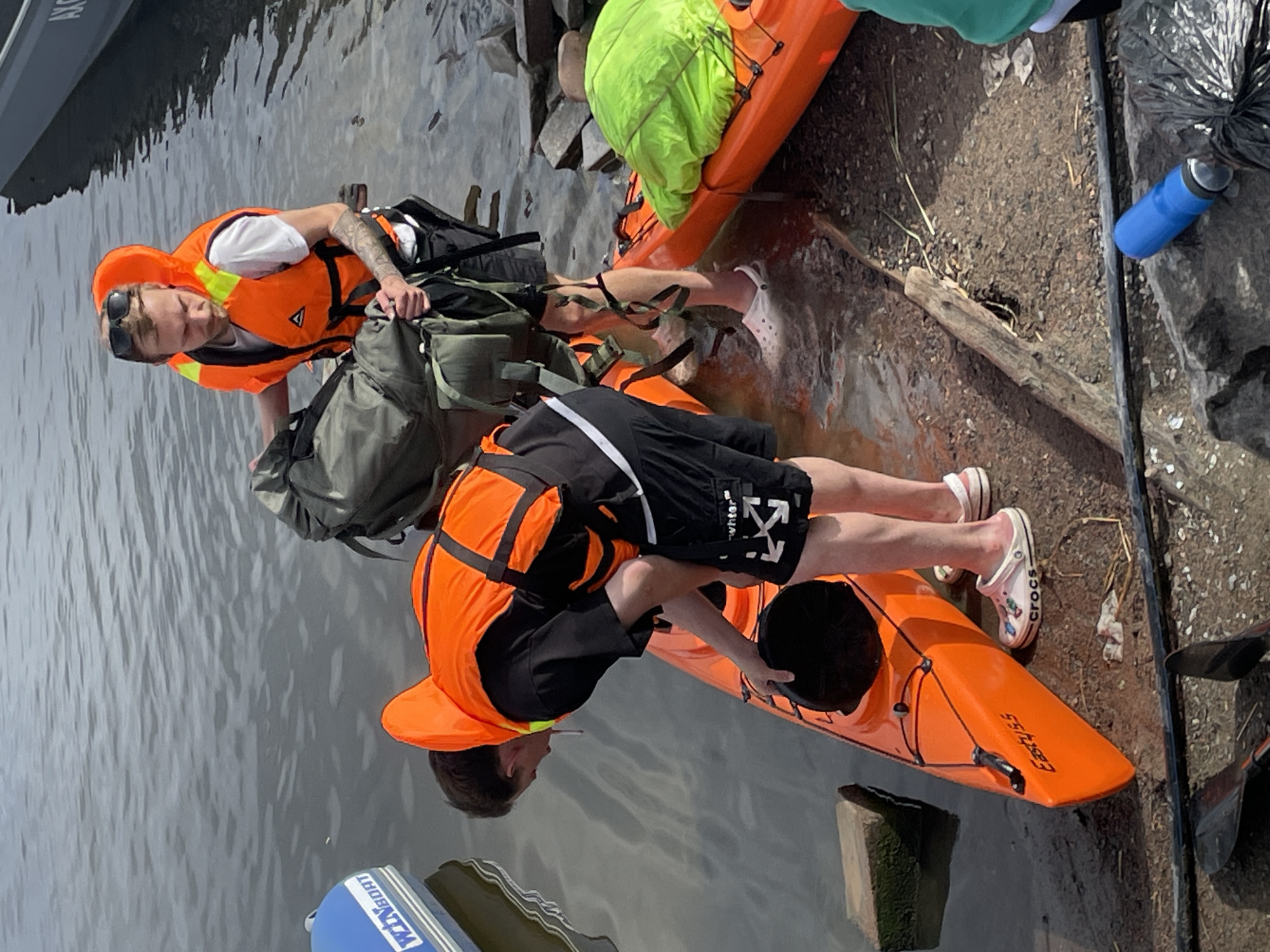 Two individuals are seen near the water's edge, preparing to launch kayaks. The person on the left, wearing an orange life vest, black t-shirt, and black shorts with white patterns, is bent over an orange kayak, placing a black object inside. The kayak has "Eastyss" printed on its side. The person on the right, also in an orange life vest and white t-shirt, is holding a large green duffel bag and appears to be assisting with loading gear. They are wearing sunglasses and have tattoos on their arm. Another orange kayak, partially visible, has a bright green bag secured on it. A blue inflatable boat with "WINBOA" visible on it is also in the water to the left. The scene takes place on a shoreline with gravel and some debris, with water in the background. The overall setting suggests an outdoor activity near a body of water.