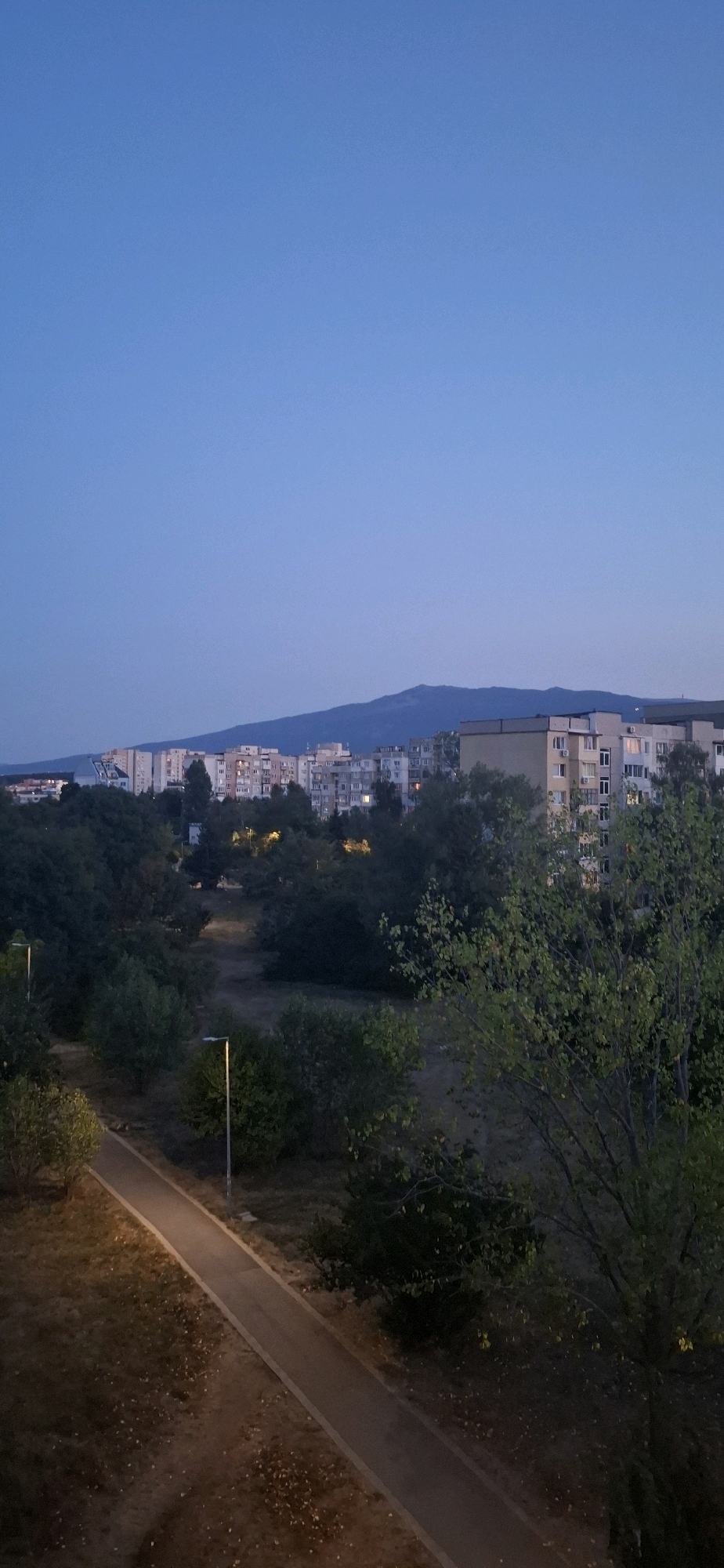 The image depicts a twilight scene in the city of Sofia, Bulgaria, likely from a residential area in a district like Mladost or Lyulin, known for their high-rise apartment blocks and surrounding greenery. In the foreground, a paved pathway curves through a park-like setting with trees and scattered fallen leaves. A lamppost illuminates a section of the path. Beyond the park, a dense collection of multi-story apartment buildings dominates the middle ground, their windows beginning to glow with interior lights, suggesting the onset of evening. In the background, a hazy mountain range, possibly Vitosha, forms a silhouette against the deep blue evening sky. The overall atmosphere is one of urban life transitioning into night, with a blend of residential infrastructure and natural elements.