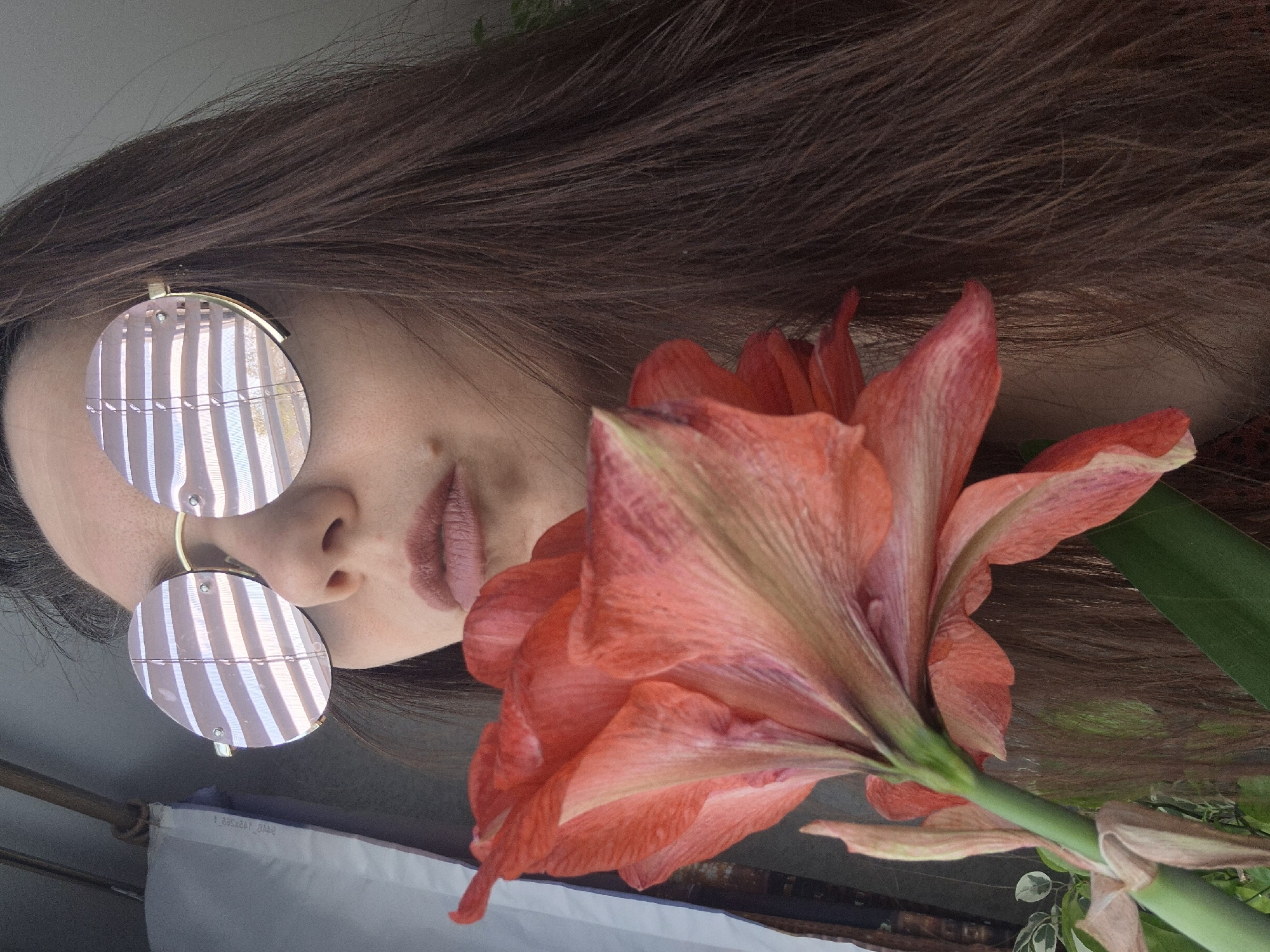 A portrait photograph, rotated counter-clockwise, showcases a person's face partially obscured by round, striped sunglasses, with an orange amaryllis flower taking prominence in the foreground. The person has long, brown hair and is wearing mauve lipstick. The flower's stem extends downwards, surrounded by green leaves and hints of other greenery. The background is blurry, suggesting an outdoor setting, but lacks specific details that would reveal the exact location in the city of Unknown, Unknown.