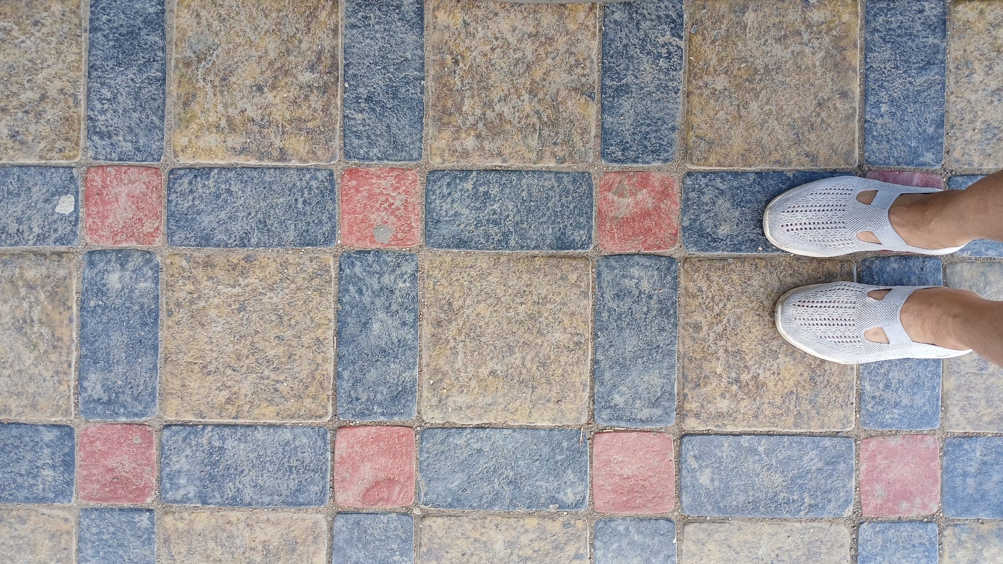 A top-down view captures a person's feet wearing light gray knit slip-on shoes standing on a patterned walkway made of rectangular pavers. The pavers are arranged in a repeating pattern of blue, beige, and red rectangles and squares. The scene is set on a paved area, possibly a pedestrian pathway or patio. The environment appears to be outdoors. The image provides no context or visual cues to determine if it is in the city of Oral, Kazakhstan.