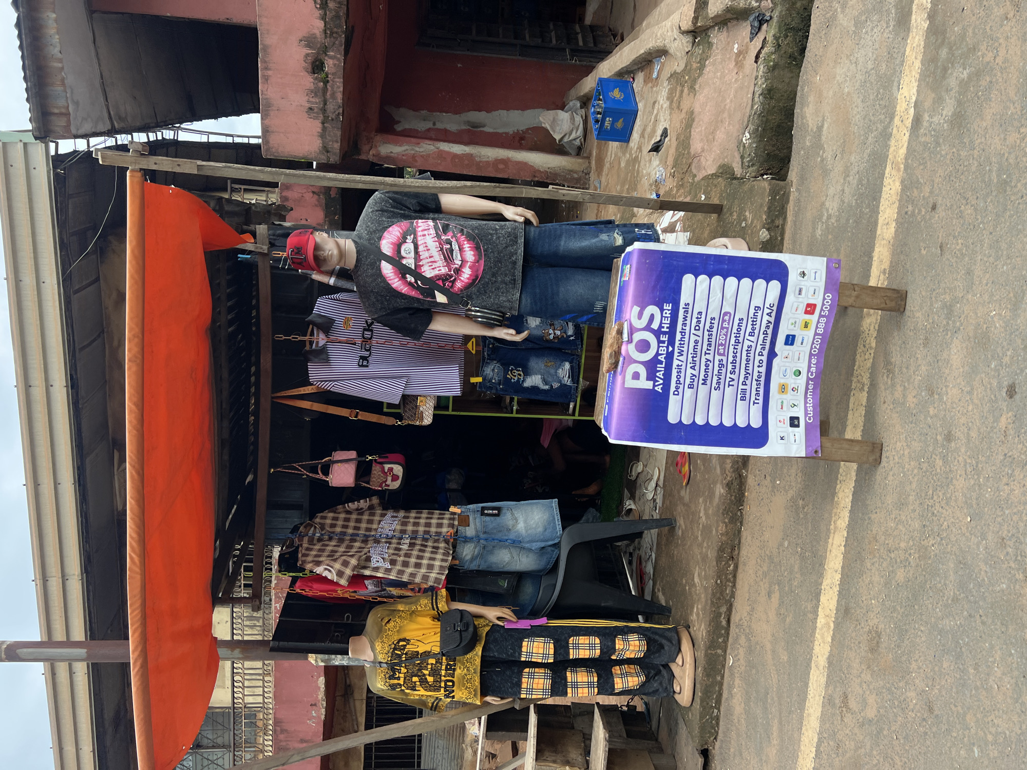 The image shows an outdoor retail stall in Nkpor, Nigeria, likely a street-side vendor. In the foreground, a purple and white sign advertising "POS AVAILABLE HERE" lists services such as "Deposit/Withdrawals," "Buy Airtime / Data," "Money Transfers," "Savings (at 20% p.a.)," "TV Subscriptions," "Bill Payments / Betting," and "Transfer to PalmPay A/C." Below these services are various logos of telecommunication and financial service providers. The sign is supported by two wooden posts.
The stall itself is covered by a bright orange awning supported by wooden beams. Displayed on mannequins are clothing items, including a yellow t-shirt with black lettering on the left mannequin, paired with black pants featuring yellow plaid patterns. Another mannequin in the center wears a striped long-sleeved shirt with the word "BLACK" prominently displayed, and a dark t-shirt with a graphic of pink lips on the right mannequin, accompanied by blue denim shorts or wide-leg pants. Several other garments, including plaid shirts and denim shorts, are hung on display racks behind the mannequins. Two small pink handbags are also displayed.
The ground in front of the stall is a paved surface with a yellow line painted on it. Scattered on the ground are flip-flops and debris. To the right of the stall, a blue crate containing what appears to be beverage bottles is visible. The background shows the facade of a pink building with corrugated iron roofing and some weathered architectural elements, suggesting a typical urban environment in Nigeria. The sky is overcast.